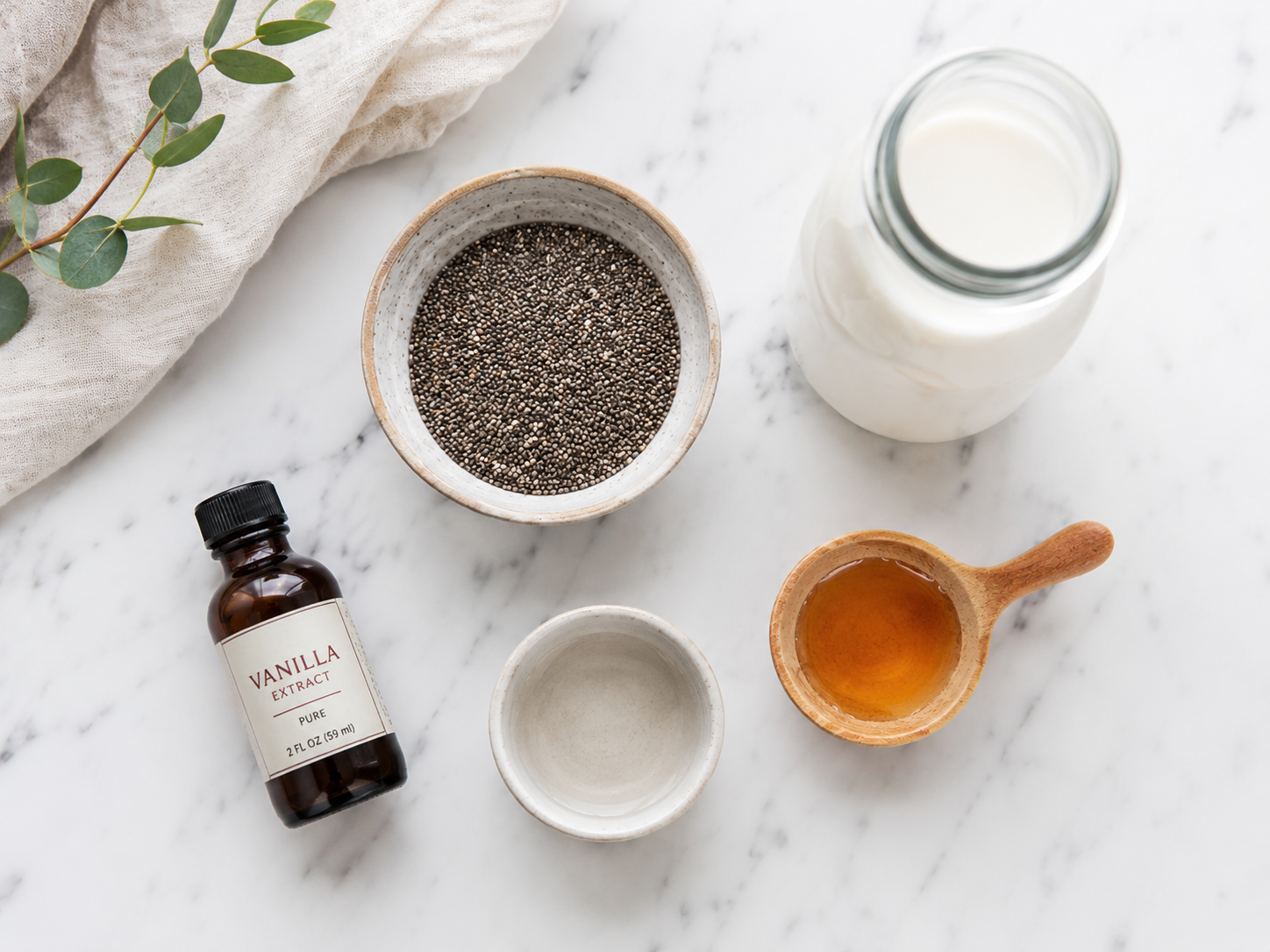 Flat lay food photography of chia pudding ingredients arranged neatly on a clean white marble countertop. A small ceramic bowl of black chia seeds, a glass bottle of milk, a small wooden spoon with golden honey, and a tiny bottle of vanilla extract. Bright, minimalist, clean aesthetic, top-down view, natural lighting