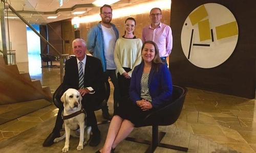 5 people in a group smiling one person is seated with a guide dog
