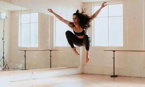 A woman with long dark hair in dance attire is jumping in a dance studio