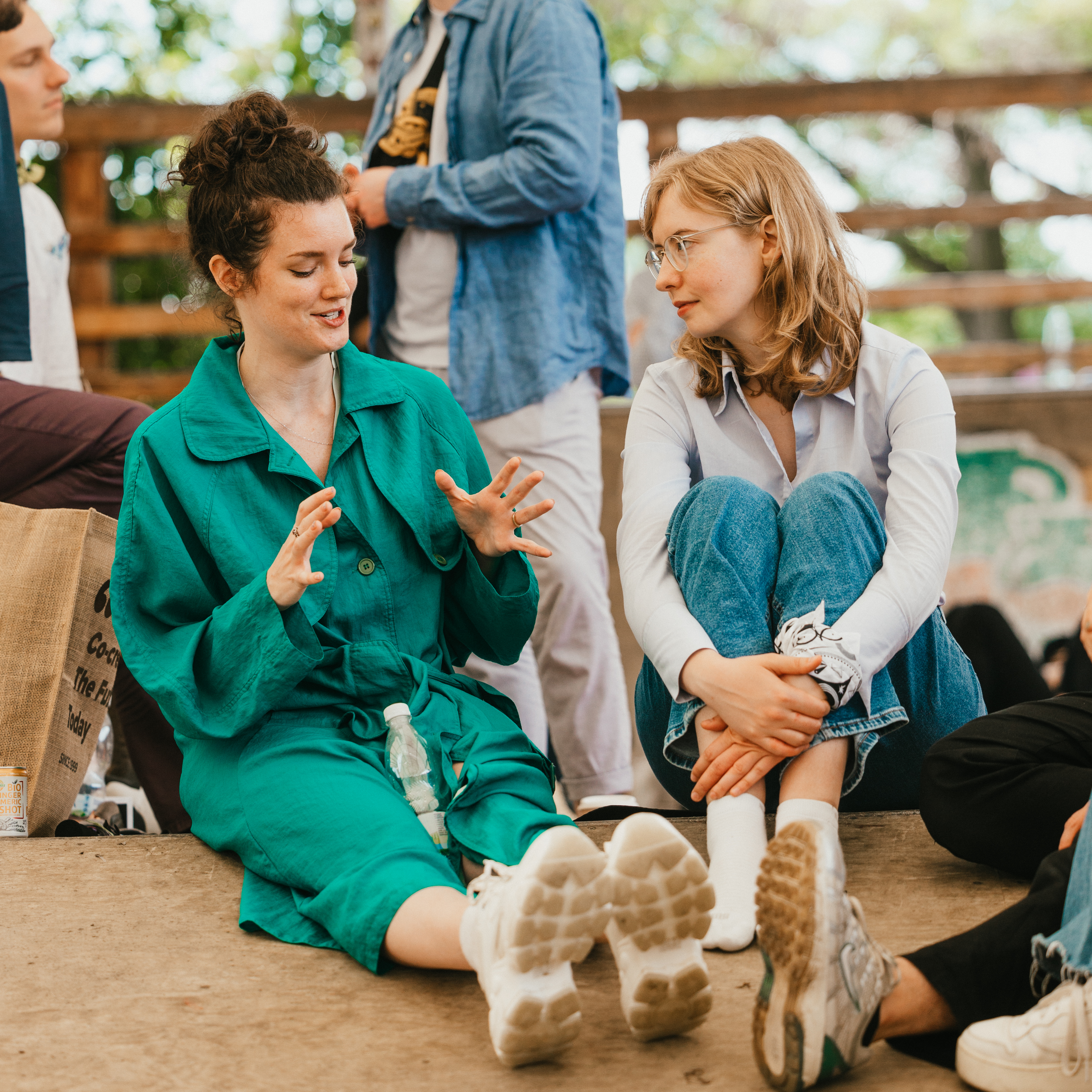 Two people sit on the ground at an outdoor event, engaged in conversation, with others nearby and greenery in the background.