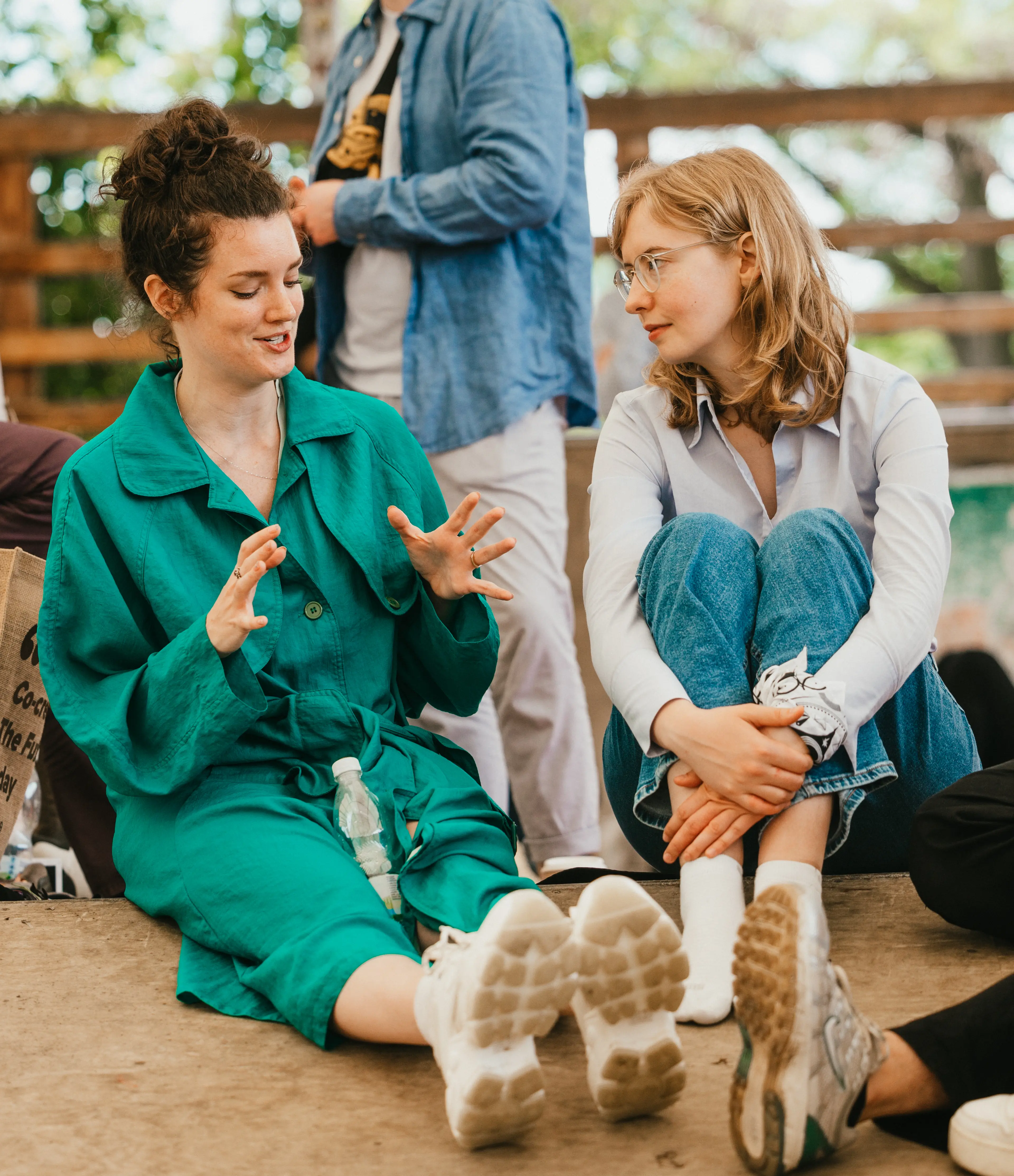 Two people sit on the ground at an outdoor event, engaged in conversation, with others nearby and greenery in the background.