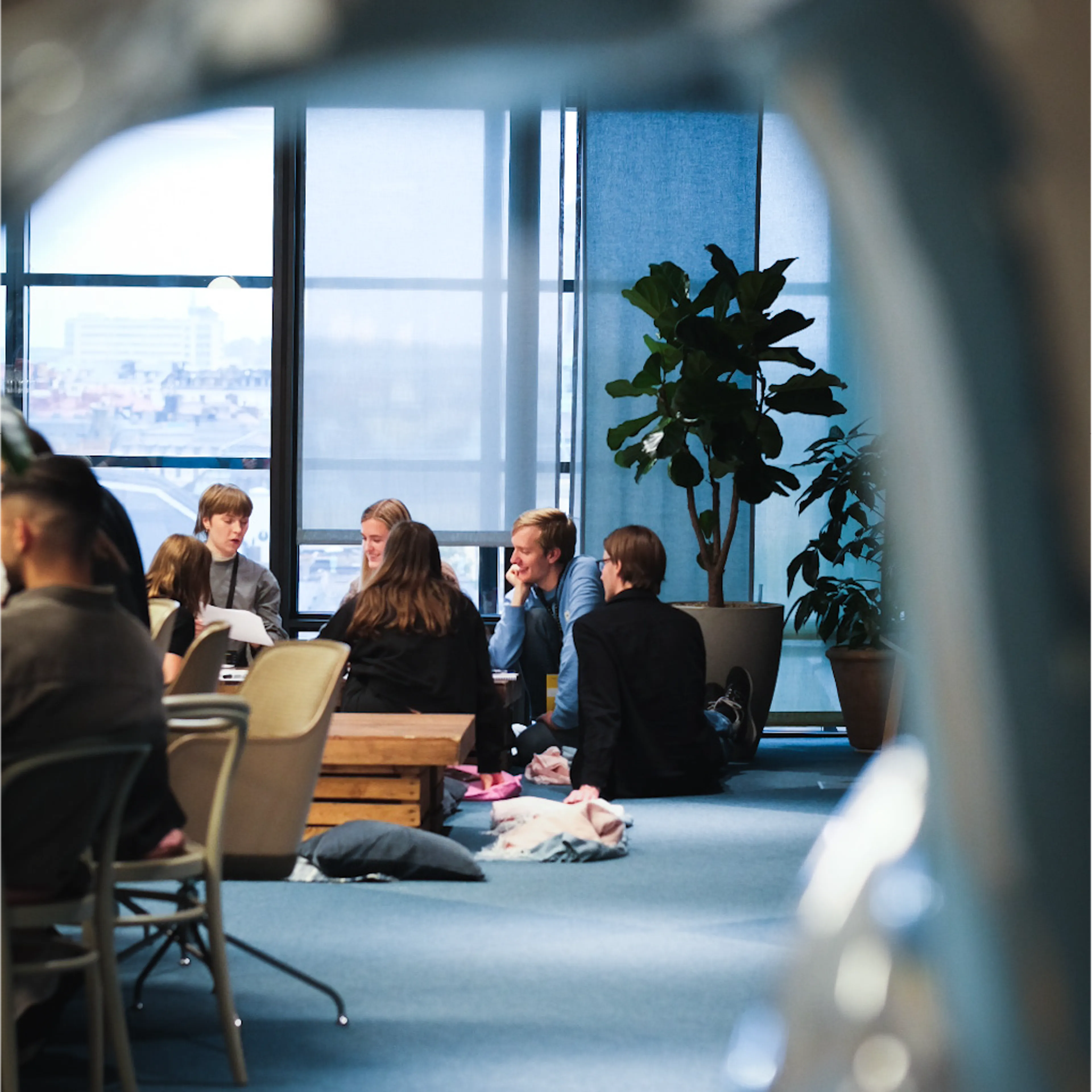 A group of people sitting on the floor in a modern office space, talking and smiling, with large windows and indoor plants in the background.