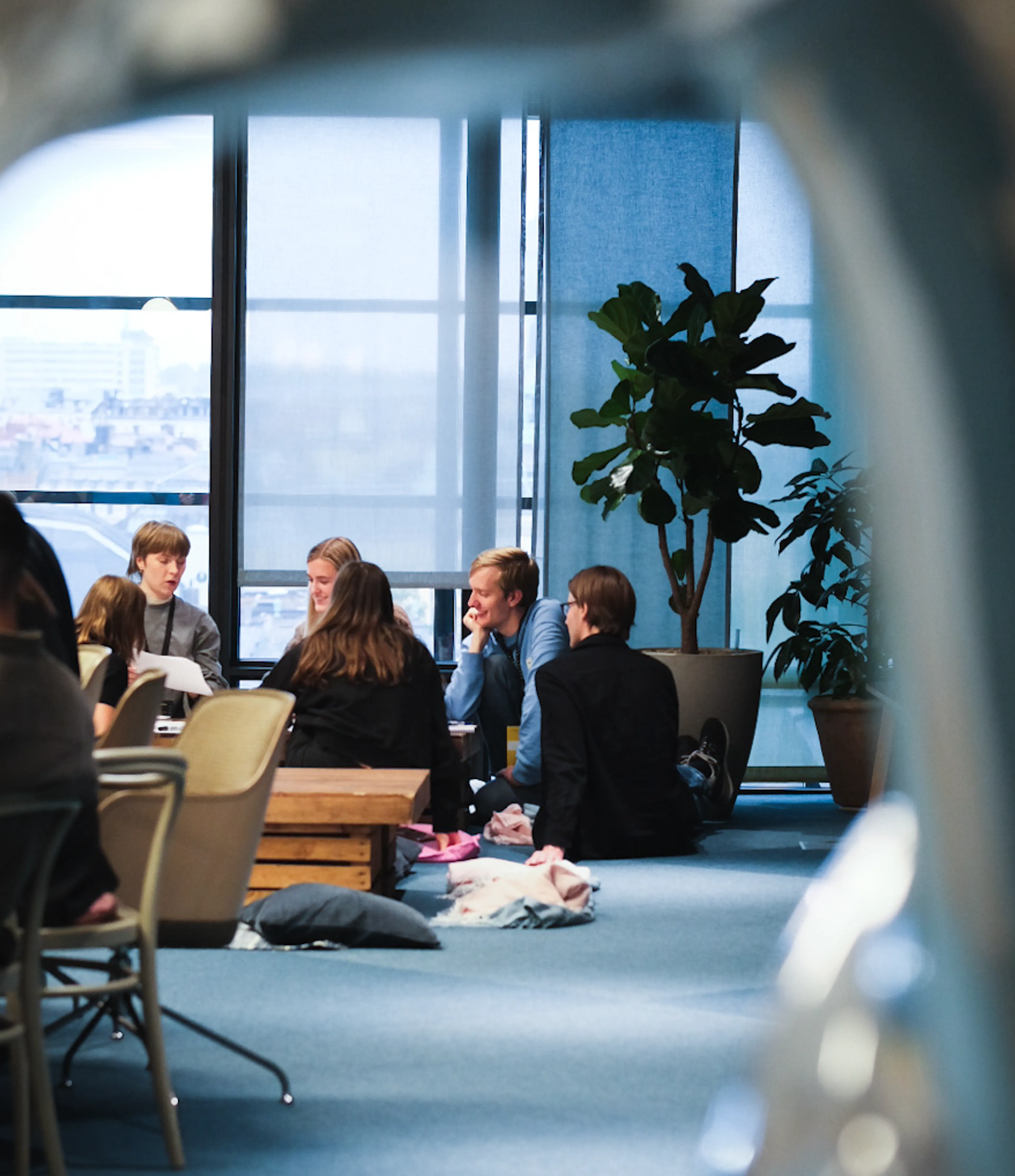 A group of people sitting on the floor in a modern office space, talking and smiling, with large windows and indoor plants in the background.