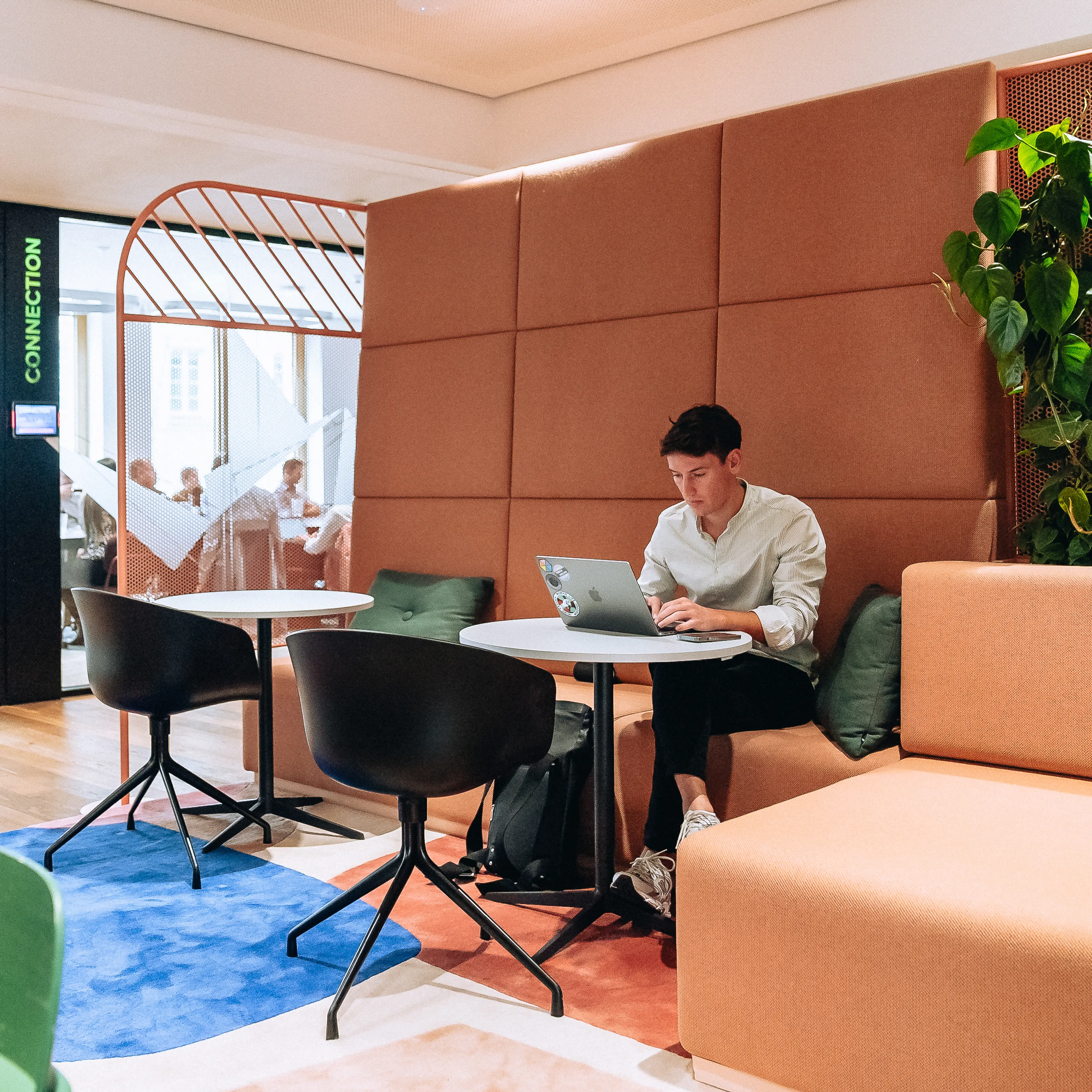 A person sits alone on an orange couch with a laptop in a brightly lit office lounge, with others visible in a nearby meeting room.