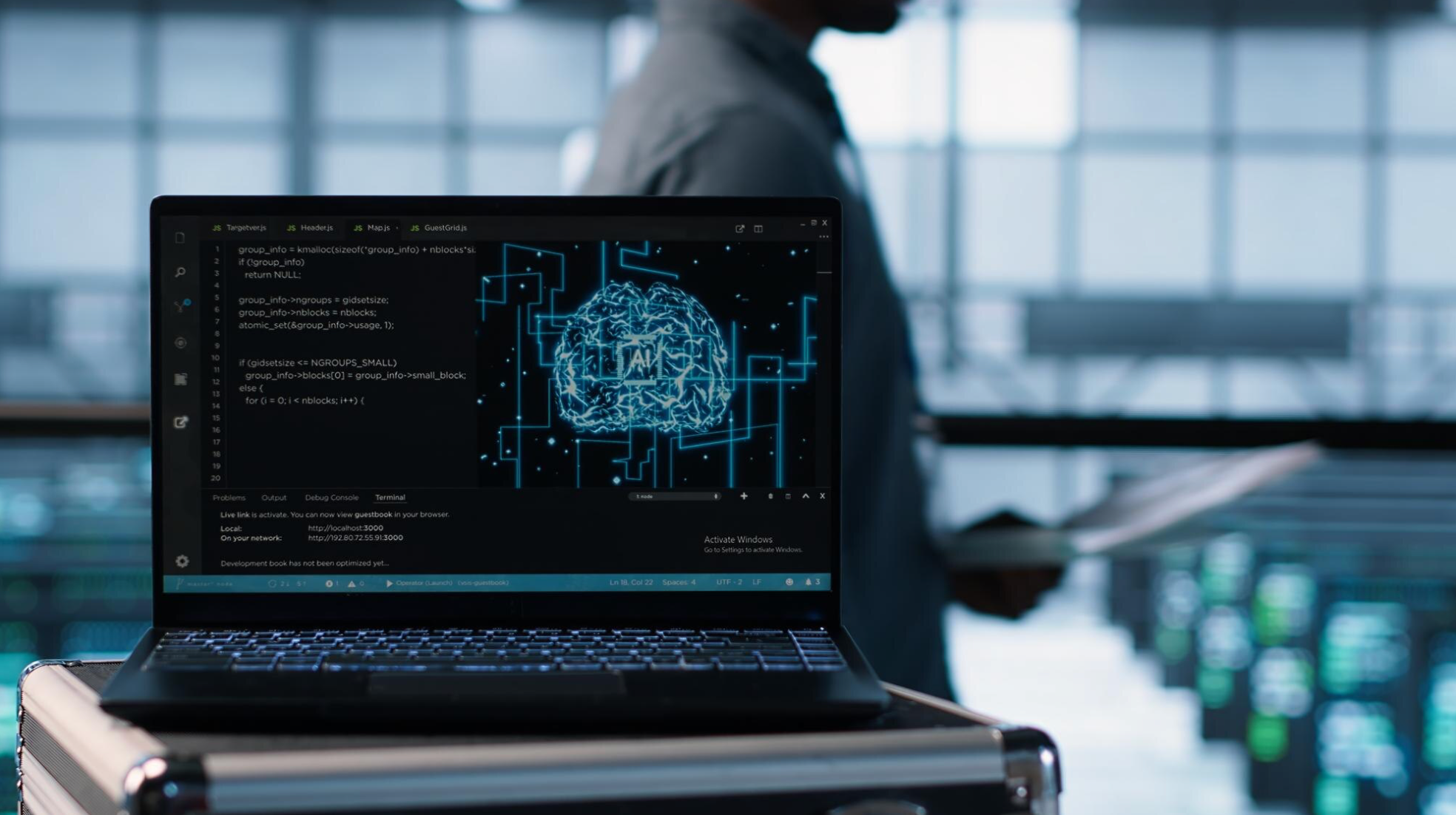 Laptop displaying artificial intelligence (AI) code and a digital brain graphic on the screen, with a blurred person holding documents and rows of servers in the background, suggesting a data center or AI development environment.