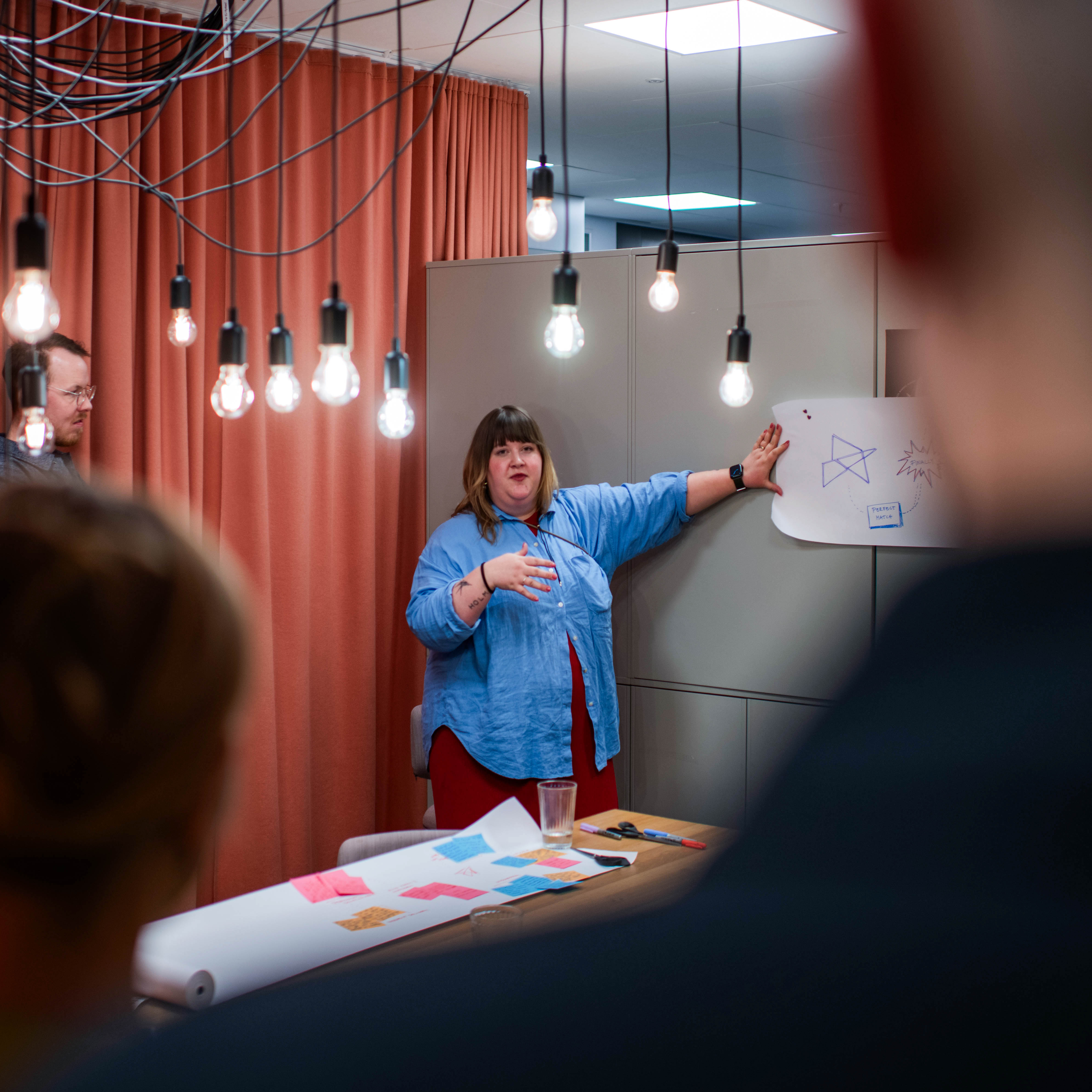 A woman stands in front of a group and a board with sketches, giving a presentation in a room with hanging light bulbs and orange curtains.