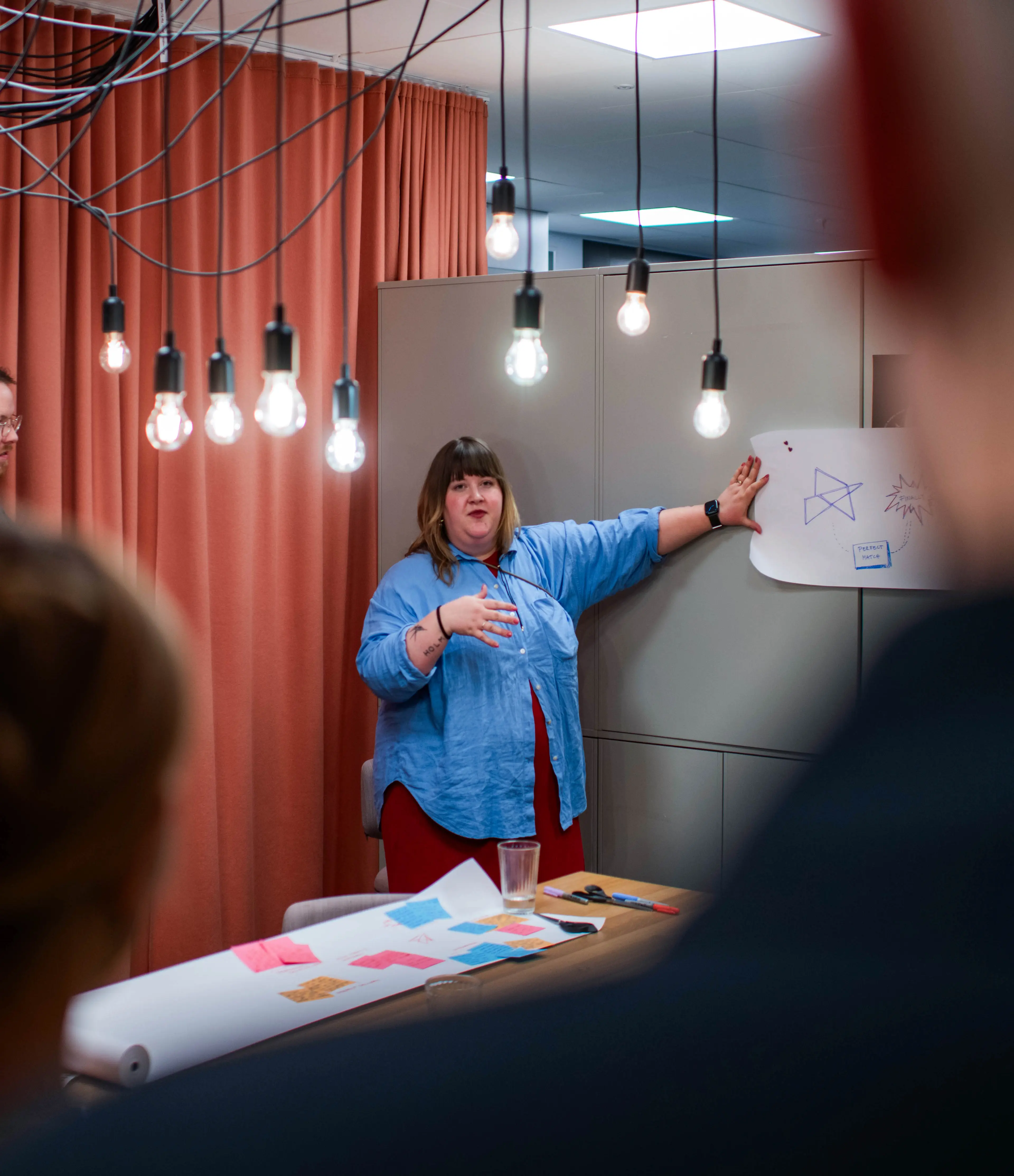 A woman stands in front of a group and a board with sketches, giving a presentation in a room with hanging light bulbs and orange curtains.
