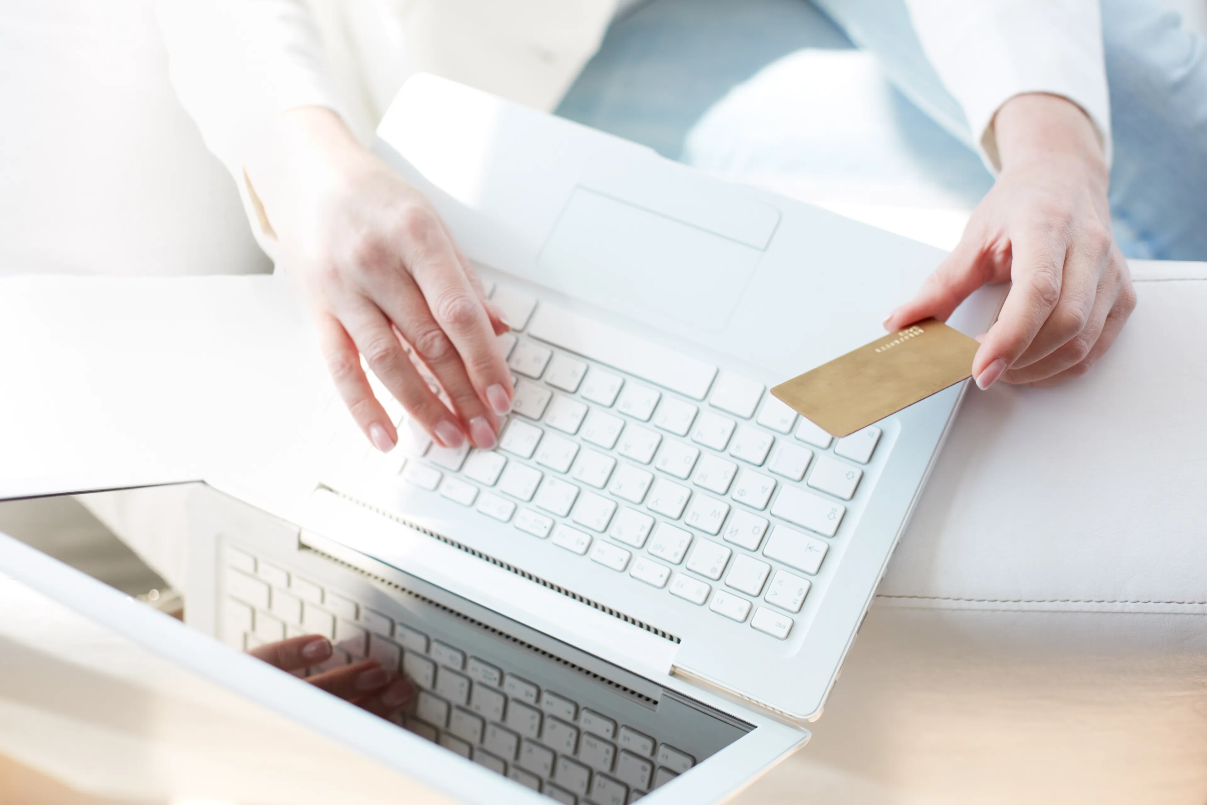 : Person holding a gold credit card in one hand while typing on a white laptop with the other hand, representing online shopping or electronic payment.