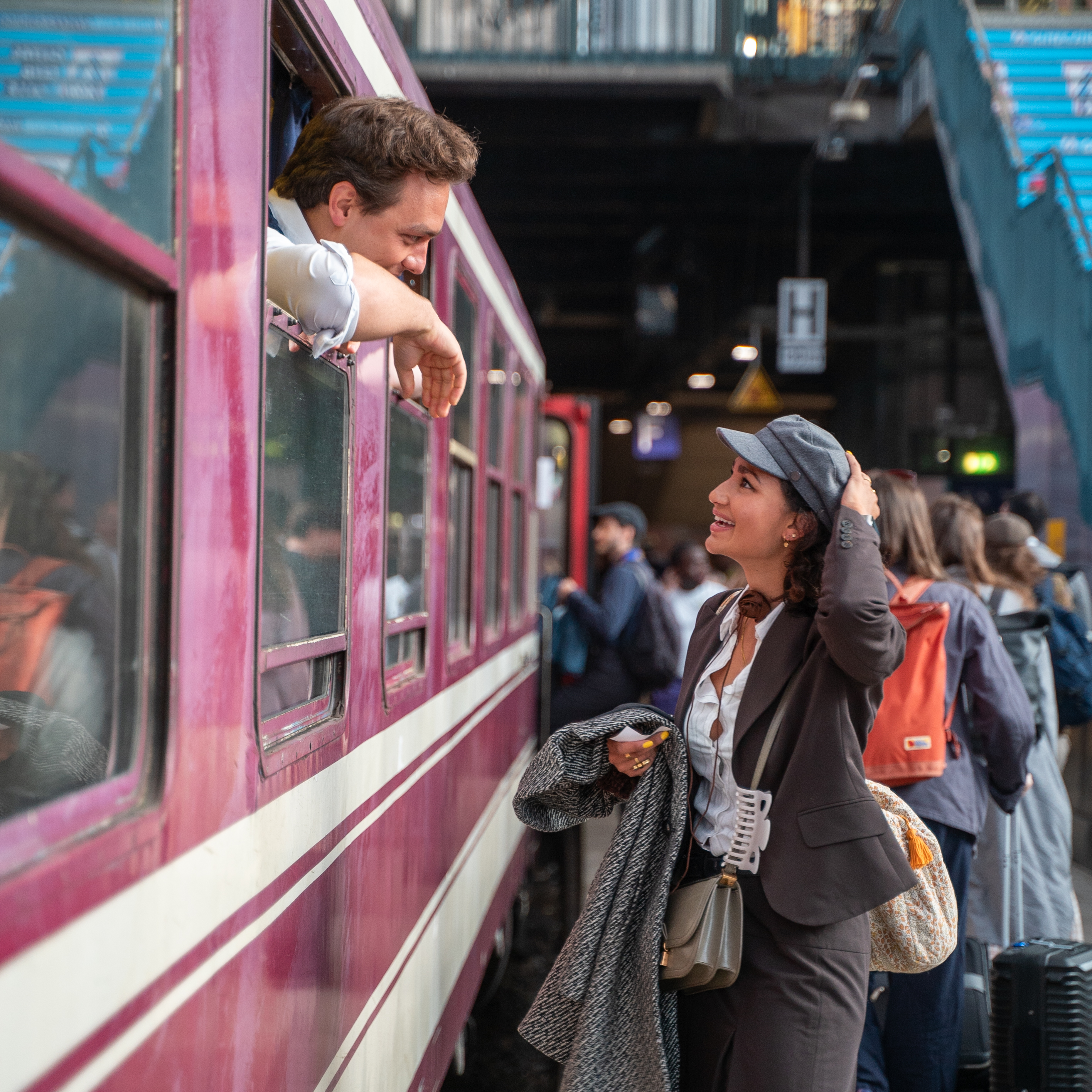 A person in a suit and hat stands on a busy train platform, smiling and talking to someone leaning out of a train window.