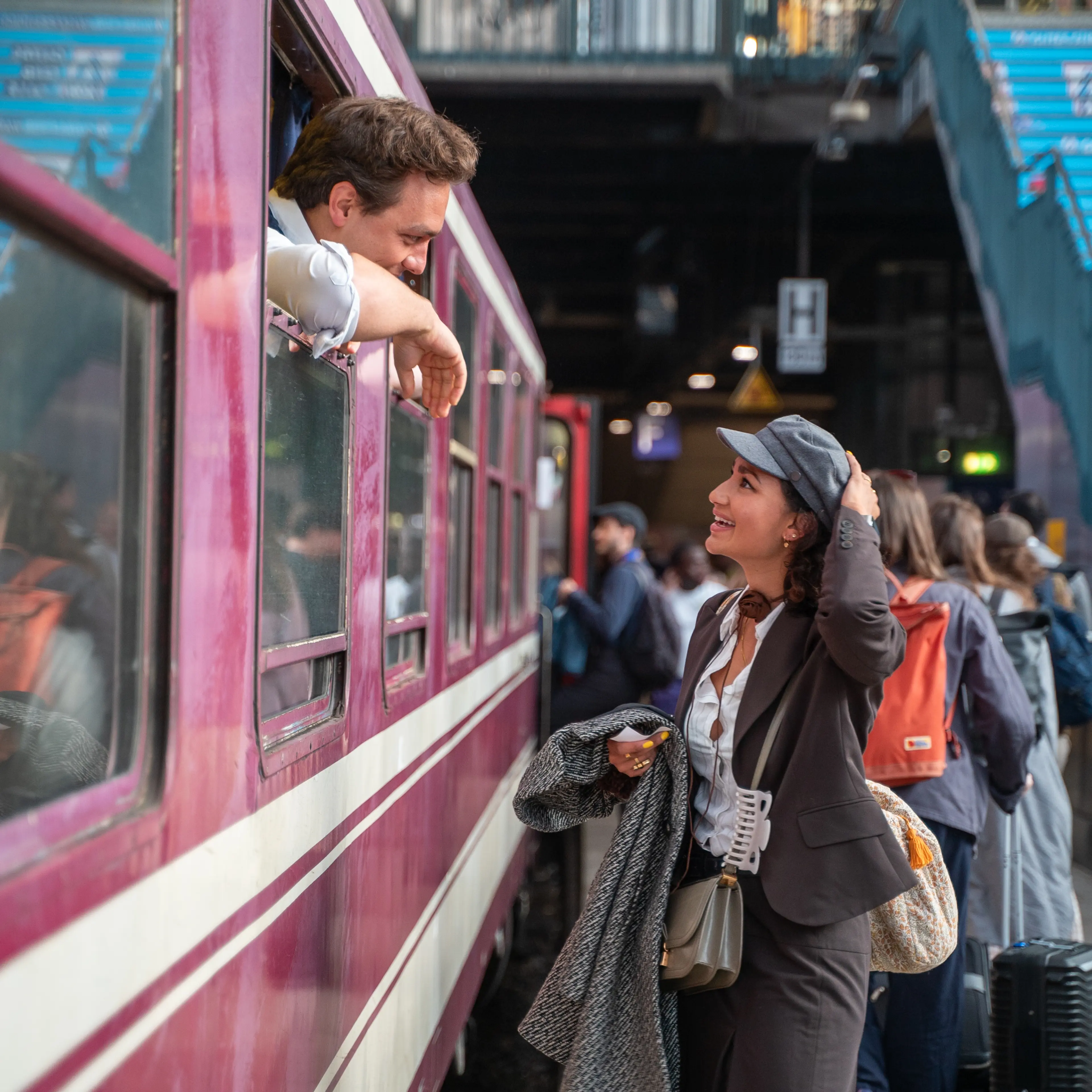 A person in a suit and hat stands on a busy train platform, smiling and talking to someone leaning out of a train window.
