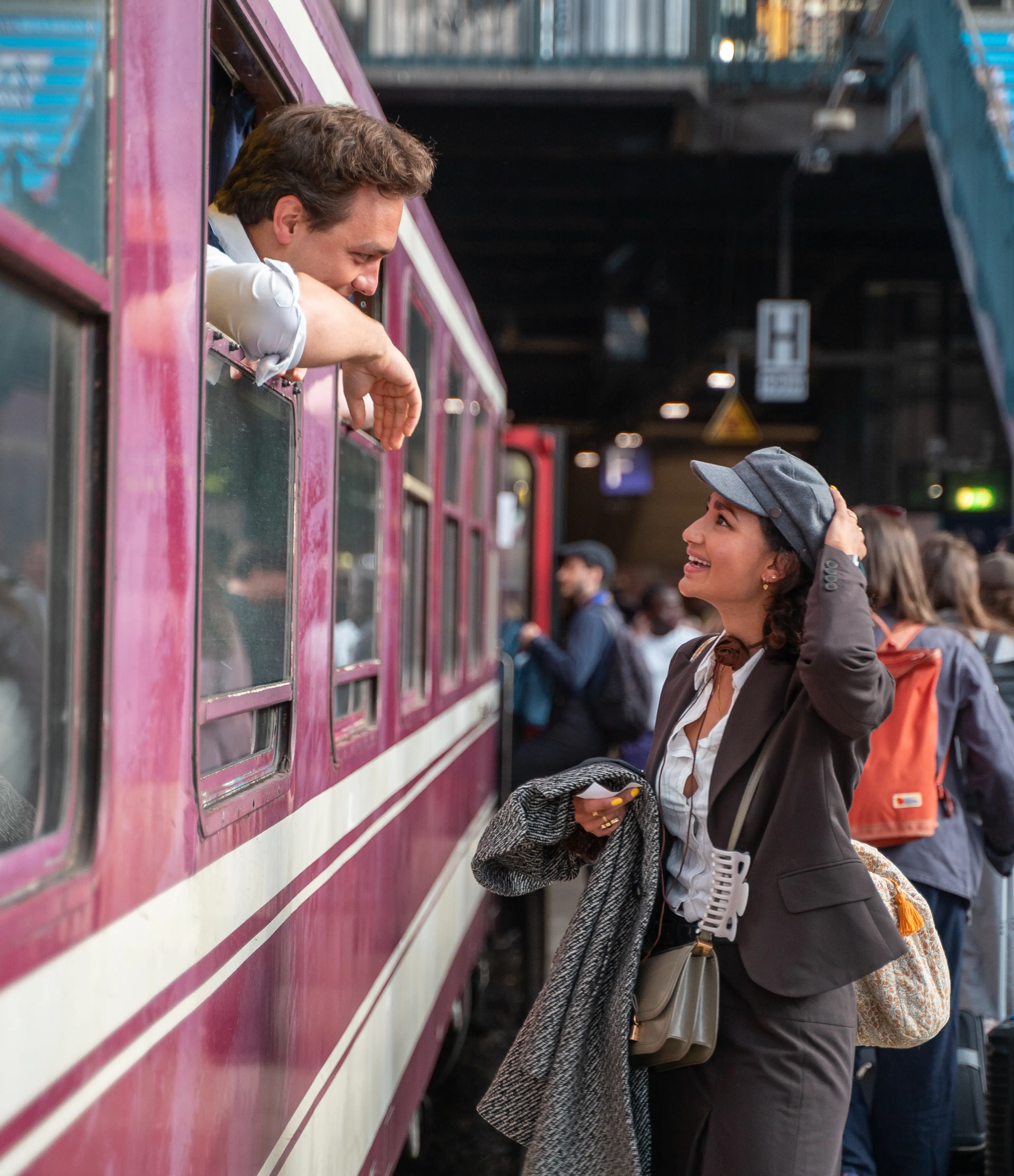 A person in a suit and hat stands on a busy train platform, smiling and talking to someone leaning out of a train window.
