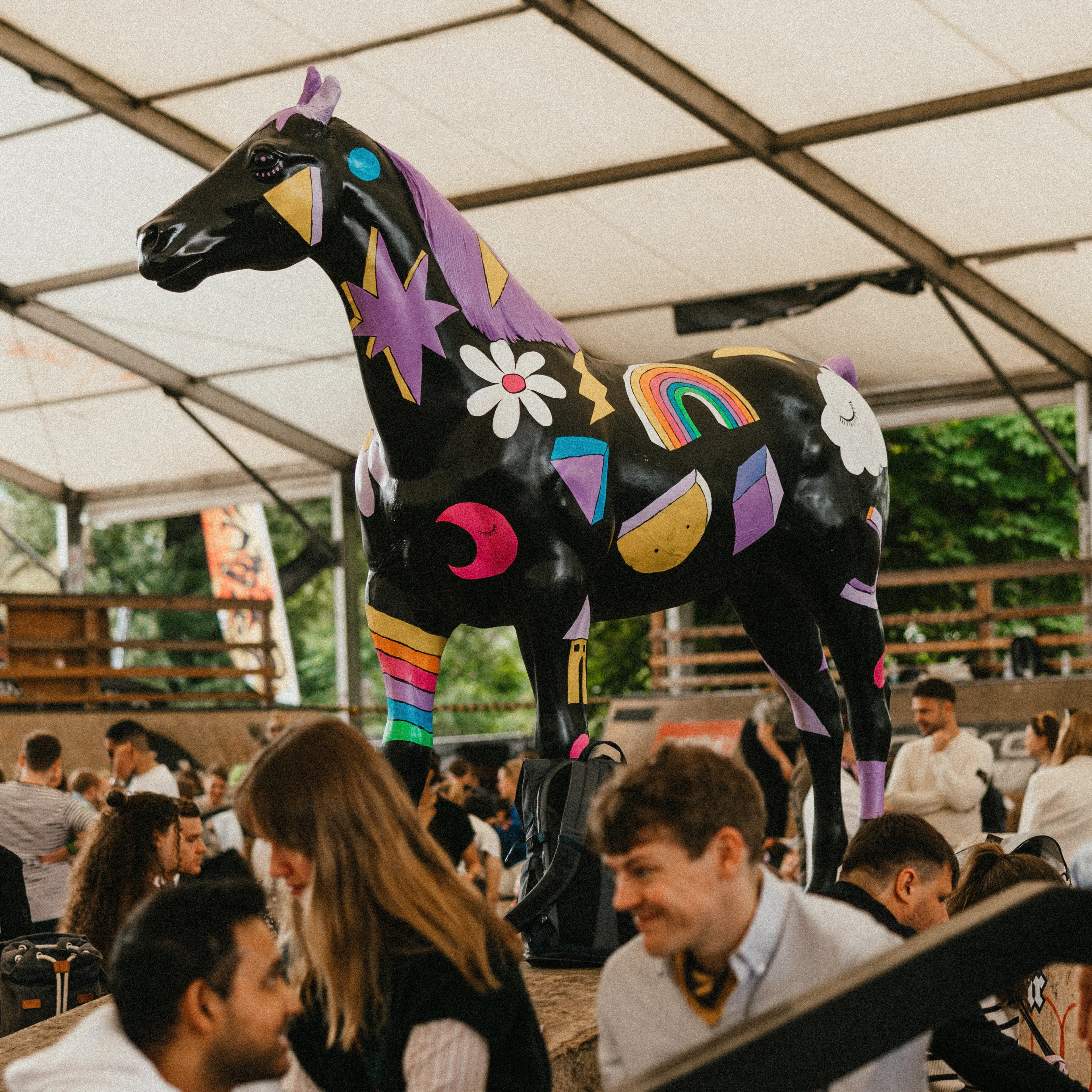 A large black horse sculpture decorated with colorful designs stands under a tent, surrounded by groups of people seated and talking.