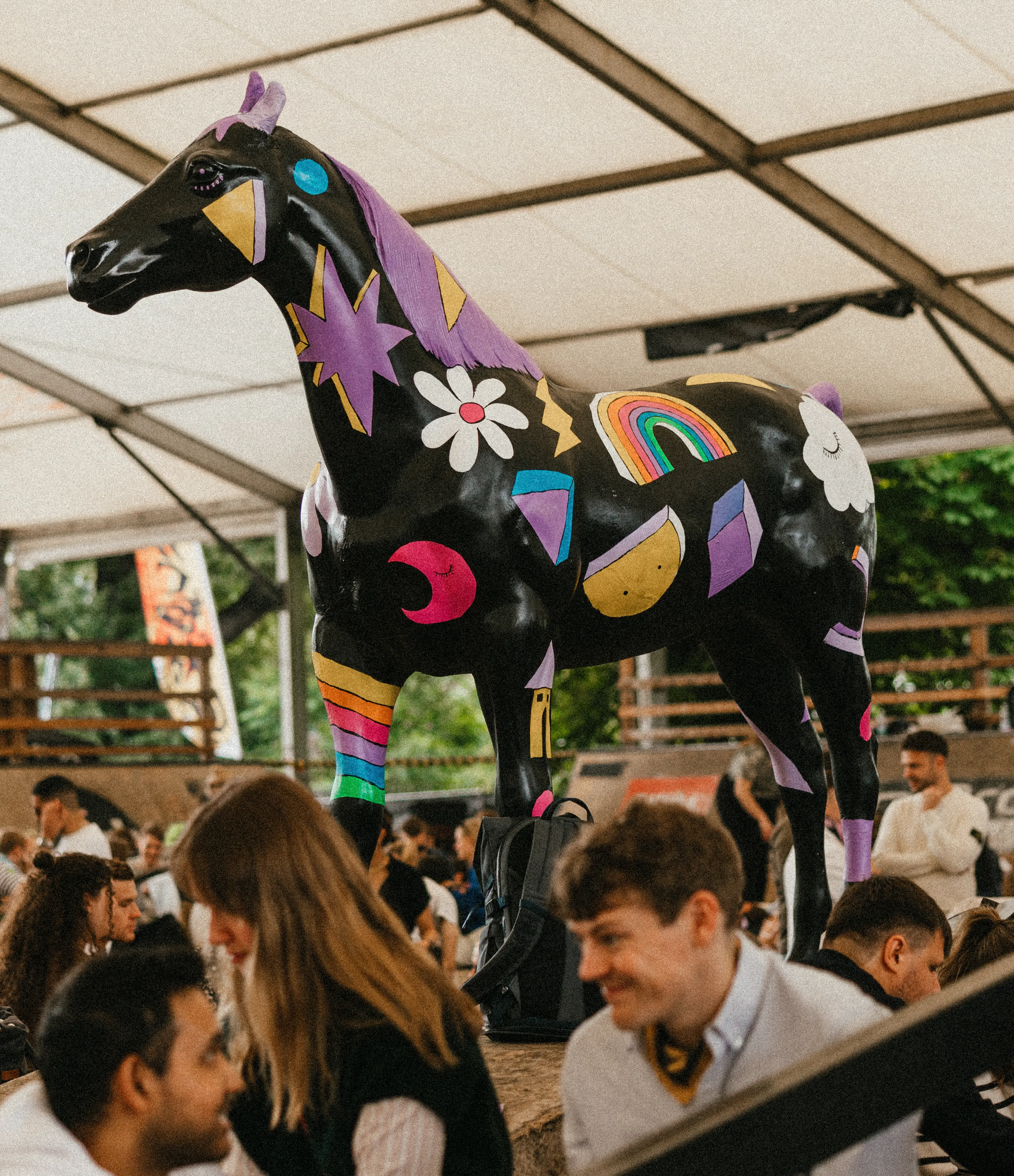 A large black horse sculpture decorated with colorful designs stands under a tent, surrounded by groups of people seated and talking.