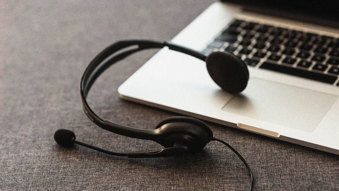 Black headset with a microphone resting on the keyboard of a silver laptop, placed on a dark surface, representing online communication or remote work