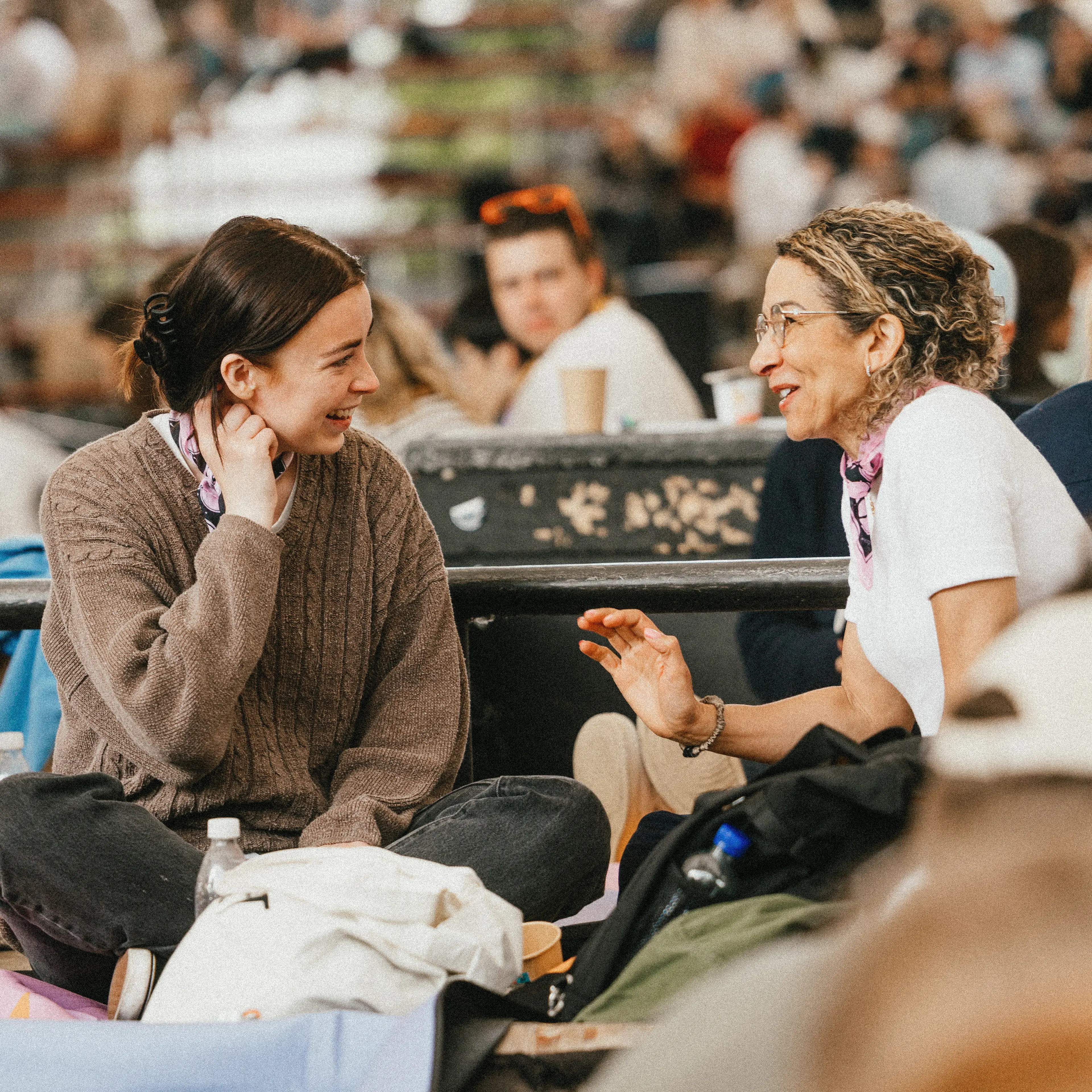 Two people sit on the ground at an outdoor event, engaged in conversation, with others nearby and greenery in the background.
