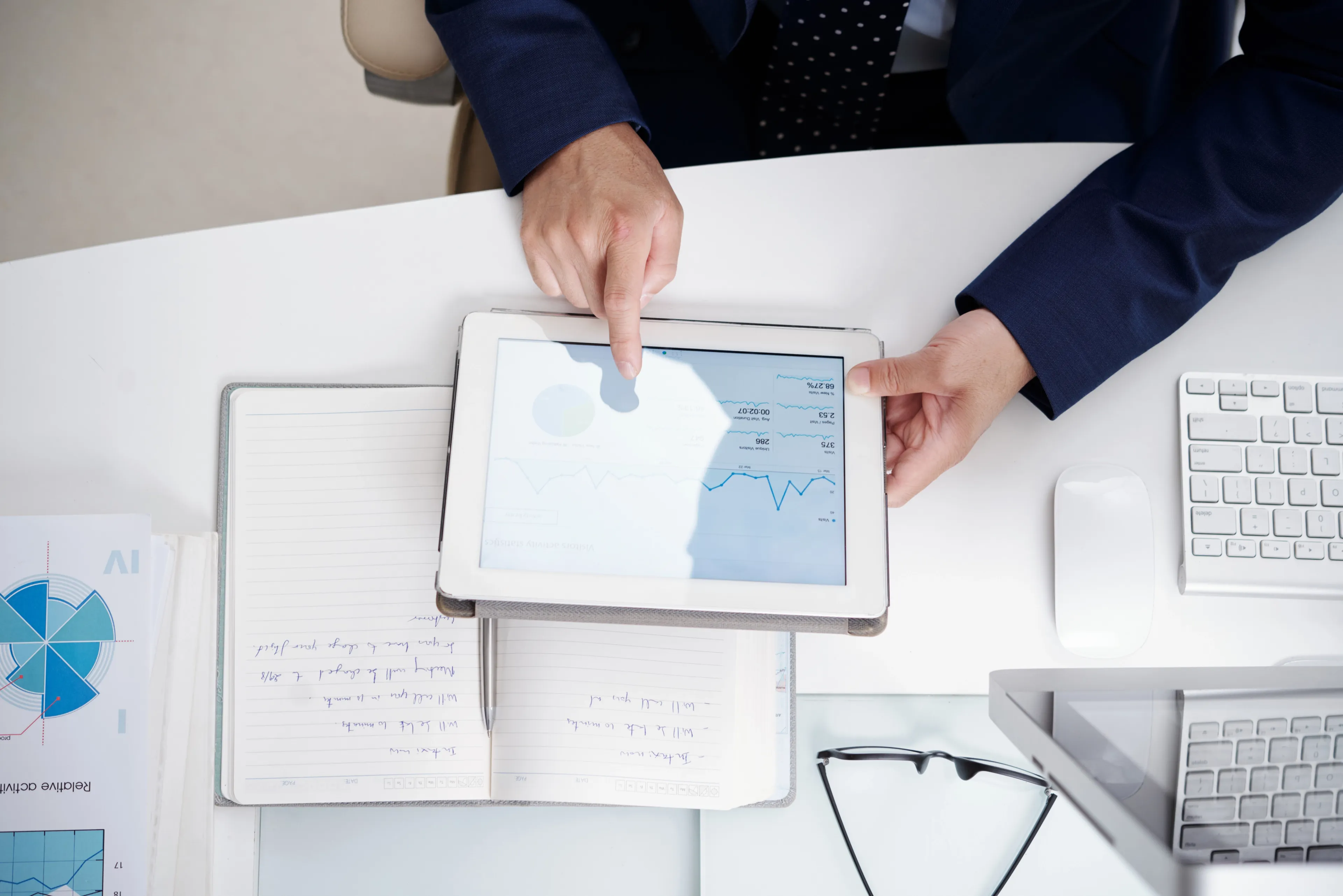 Person in business attire reviewing financial charts and graphs on a tablet, with an open notebook, keyboard, and documents on the desk in an office setting.