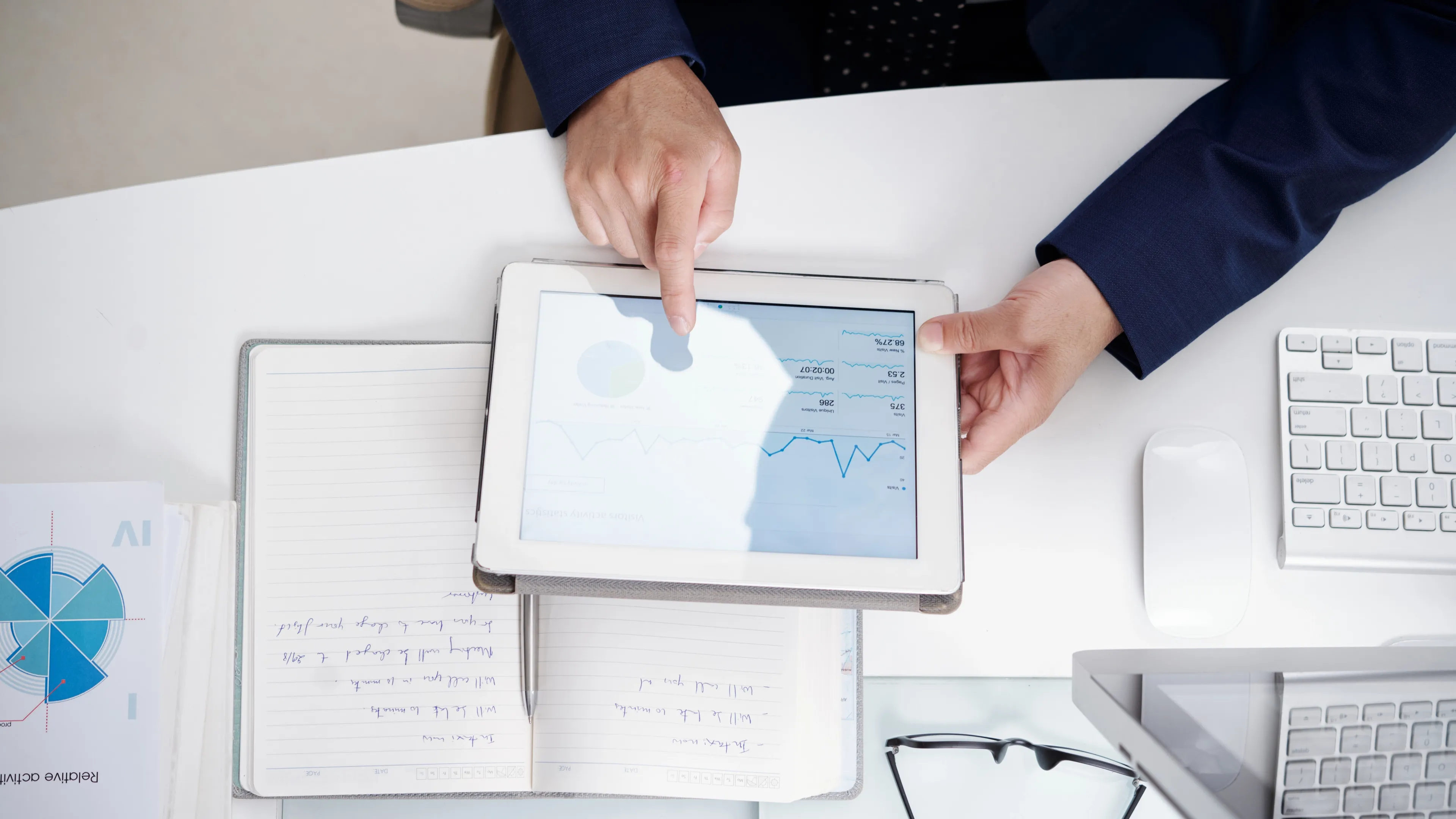 Person in business attire reviewing financial charts and graphs on a tablet, with an open notebook, keyboard, and documents on the desk in an office setting.