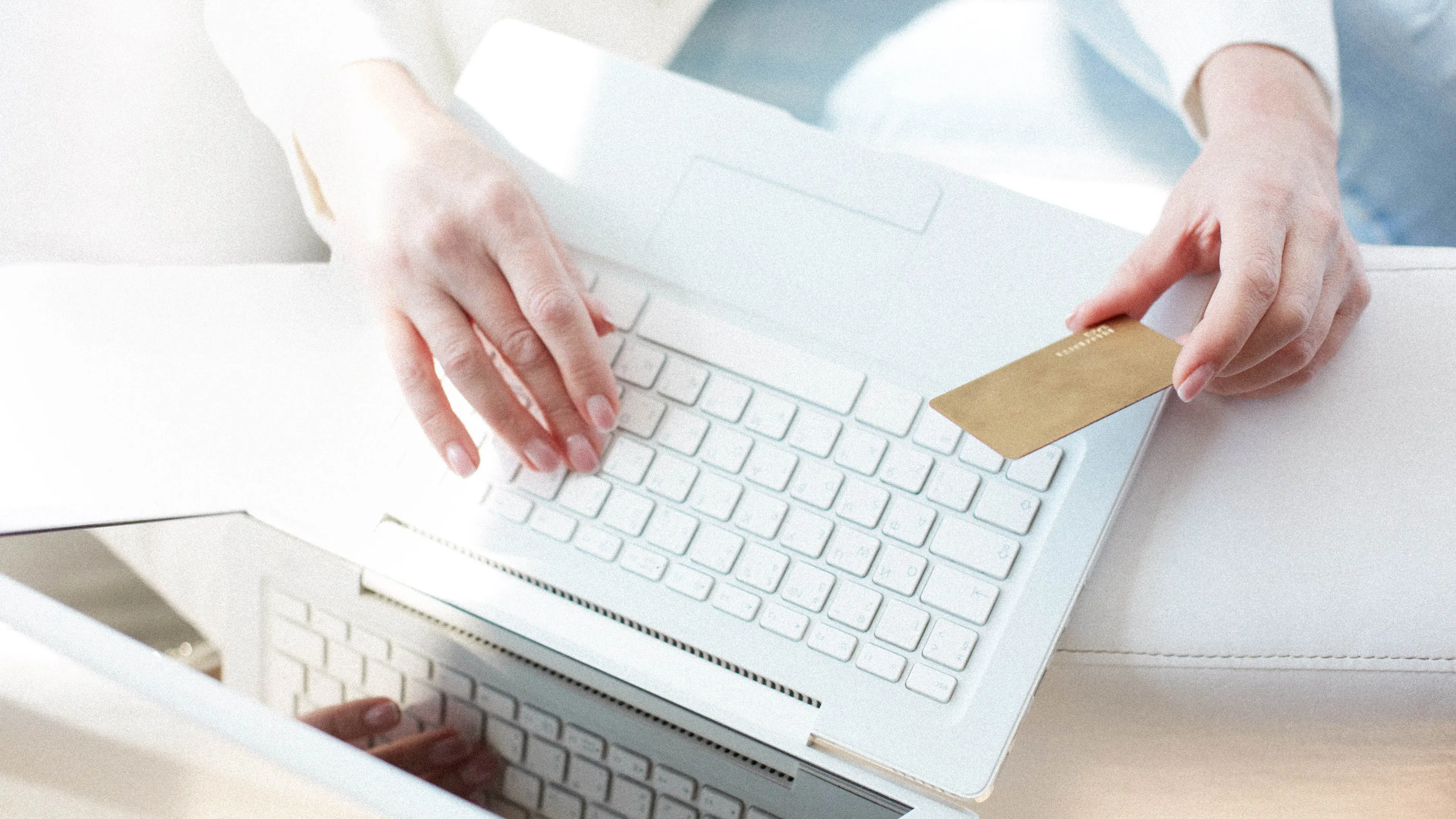 Person holding a gold credit card in one hand while typing on a white laptop with the other hand, representing online shopping or electronic payment.