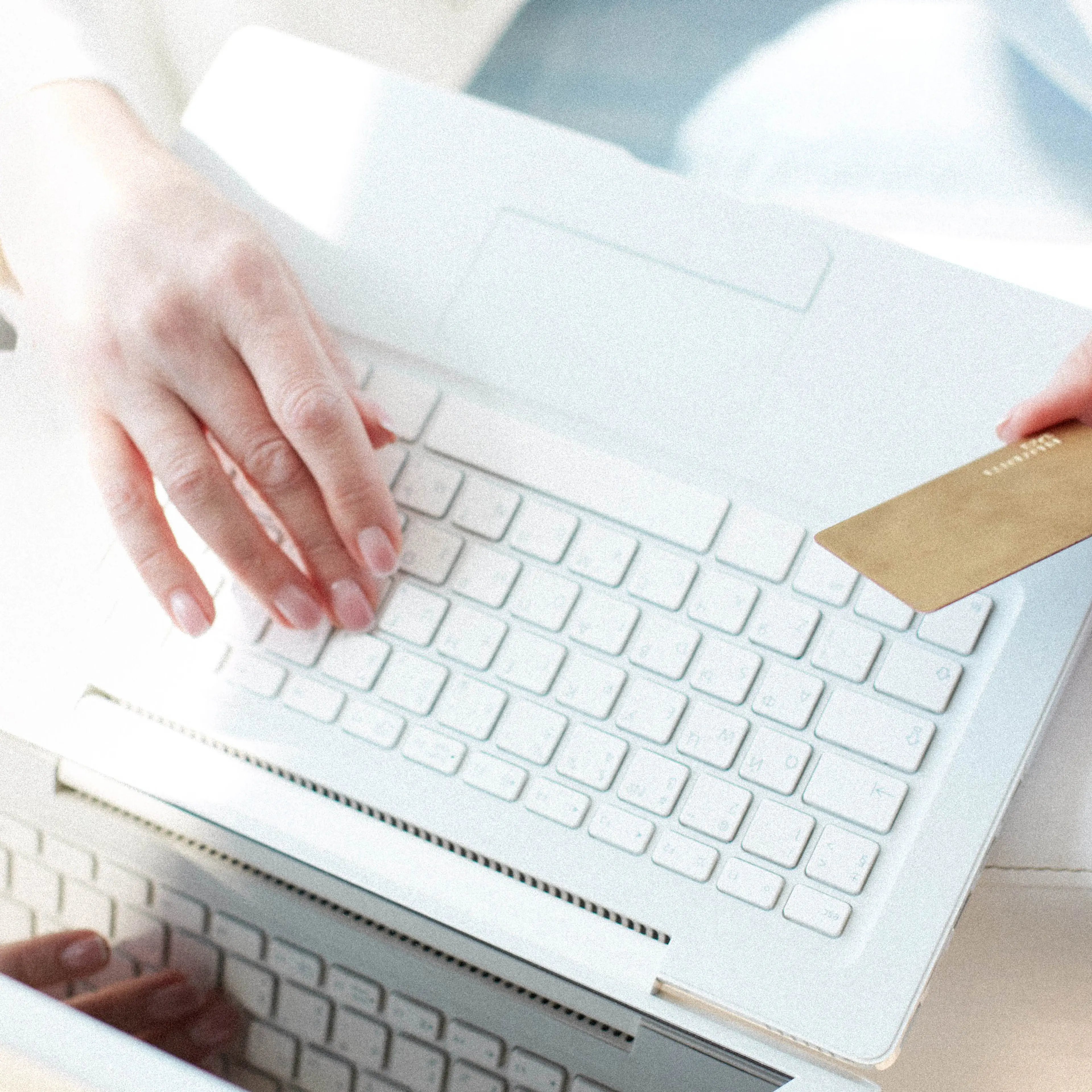 Person holding a gold credit card in one hand while typing on a white laptop with the other hand, representing online shopping or electronic payment.