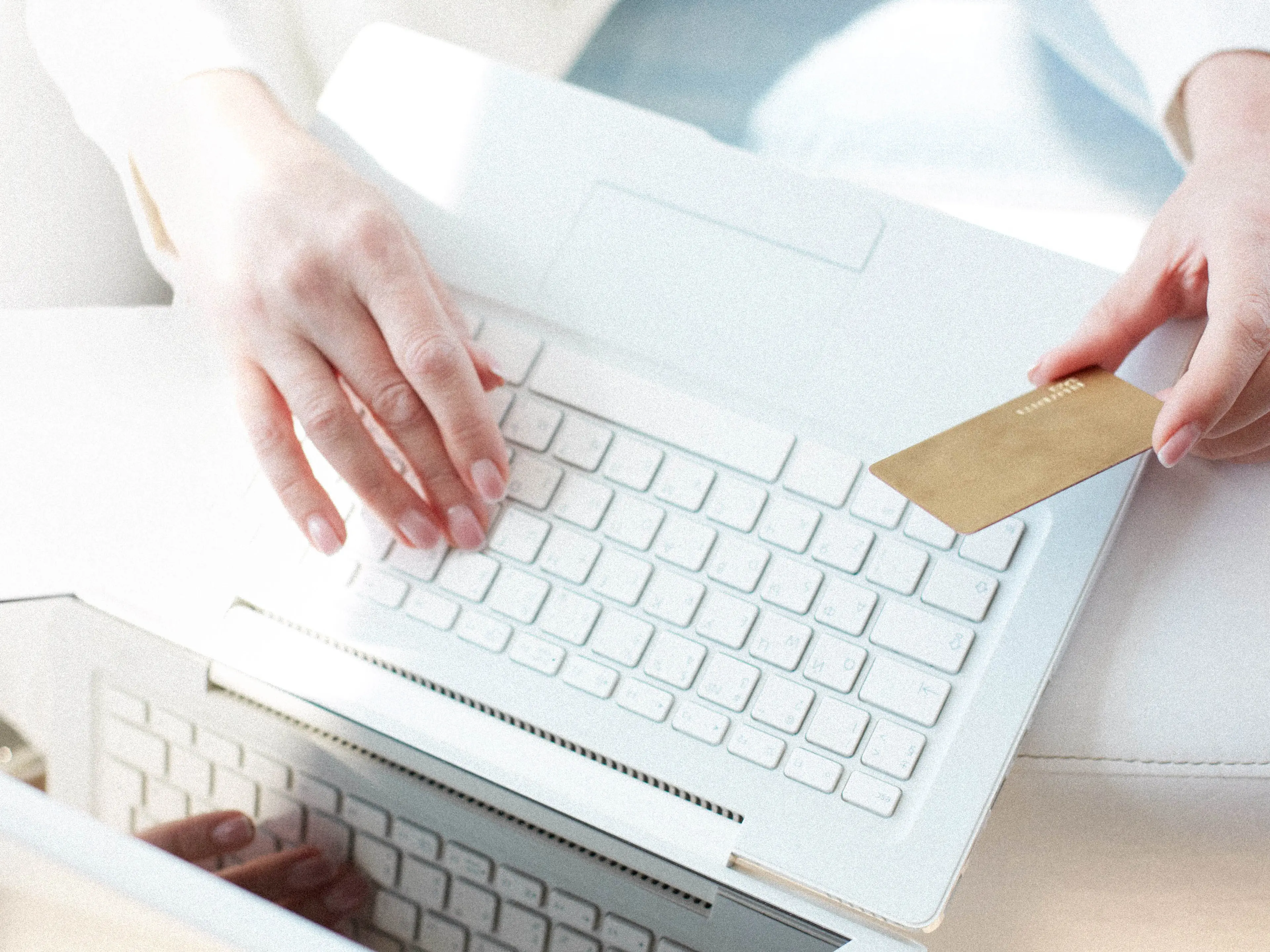 Person holding a gold credit card in one hand while typing on a white laptop with the other hand, representing online shopping or electronic payment.