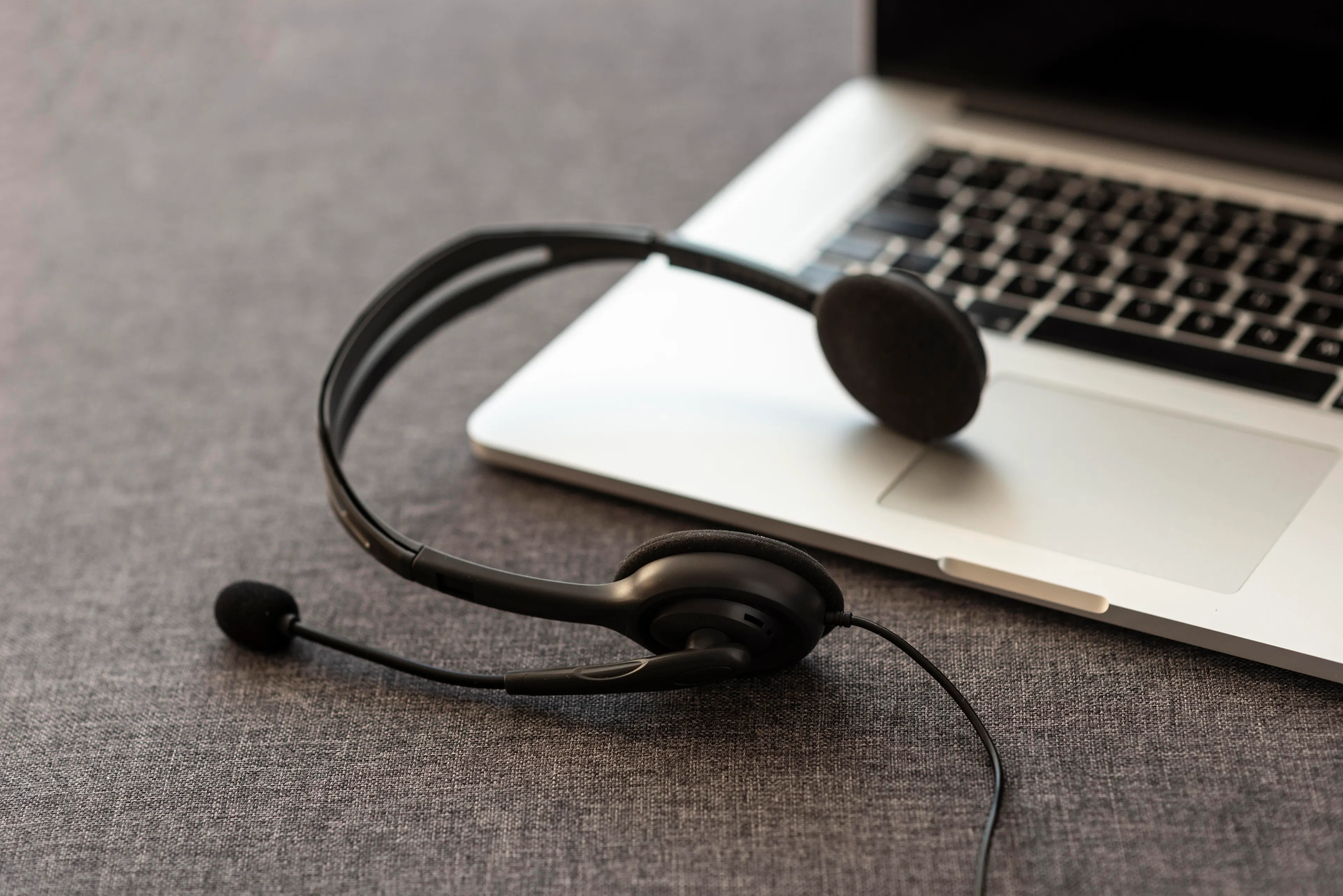 A black headset with a microphone resting on a gray surface next to an open laptop. The image suggests remote work, online communication, or customer service support.