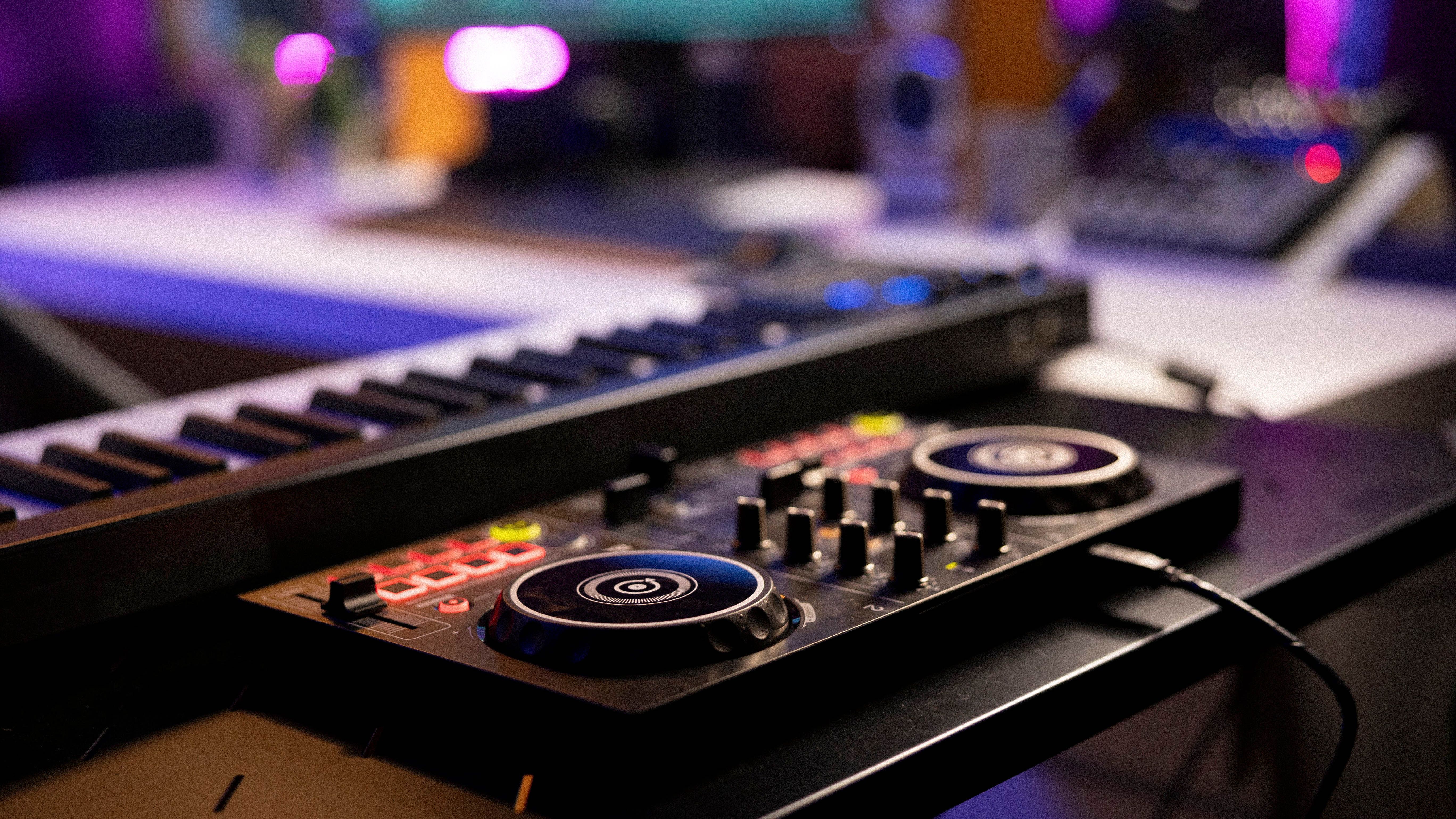 Close-up view of a DJ controller and a keyboard synthesizer on a desk in a music studio, with colorful lights and audio equipment in the background.