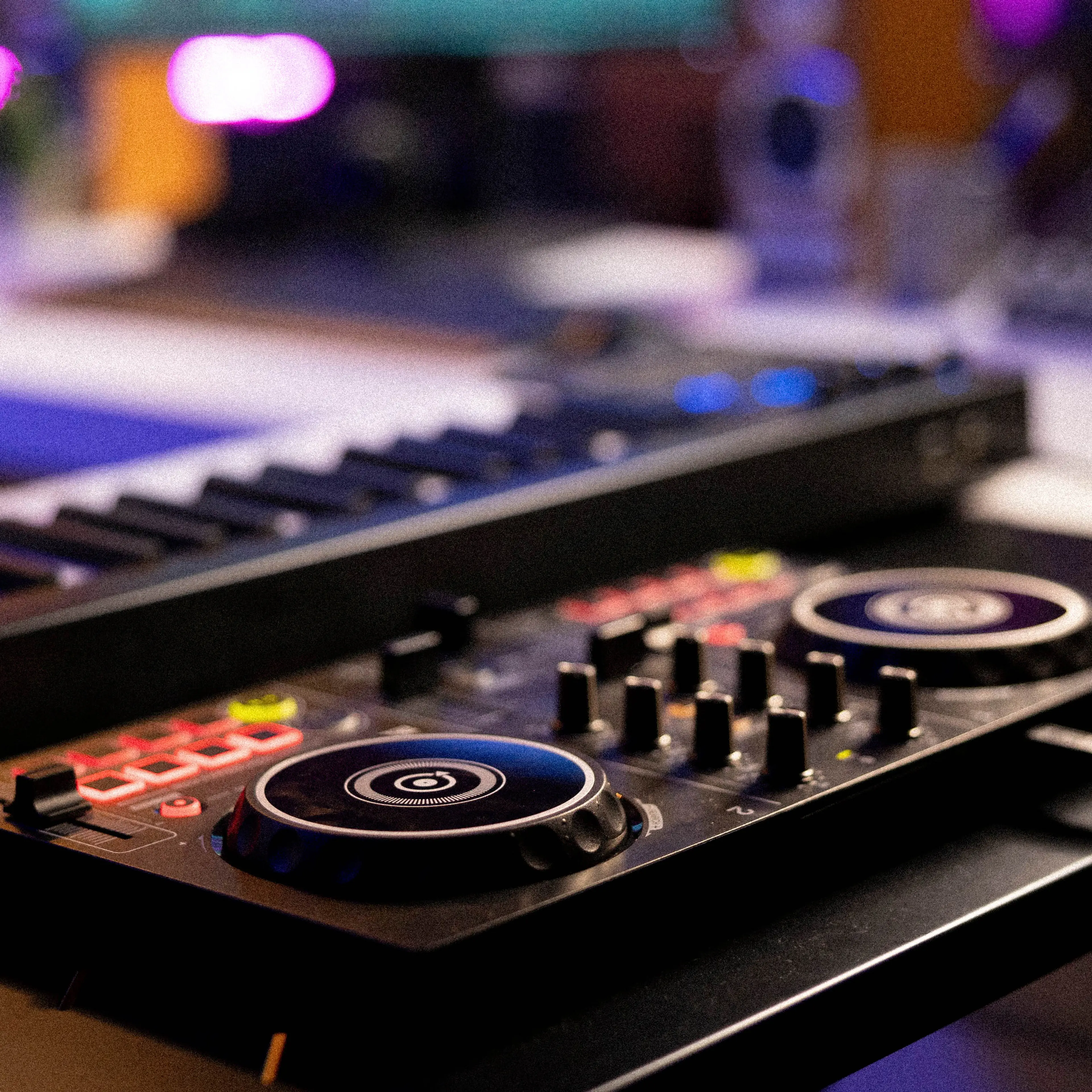 Close-up view of a DJ controller and a keyboard synthesizer on a desk in a music studio, with colorful lights and audio equipment in the background.