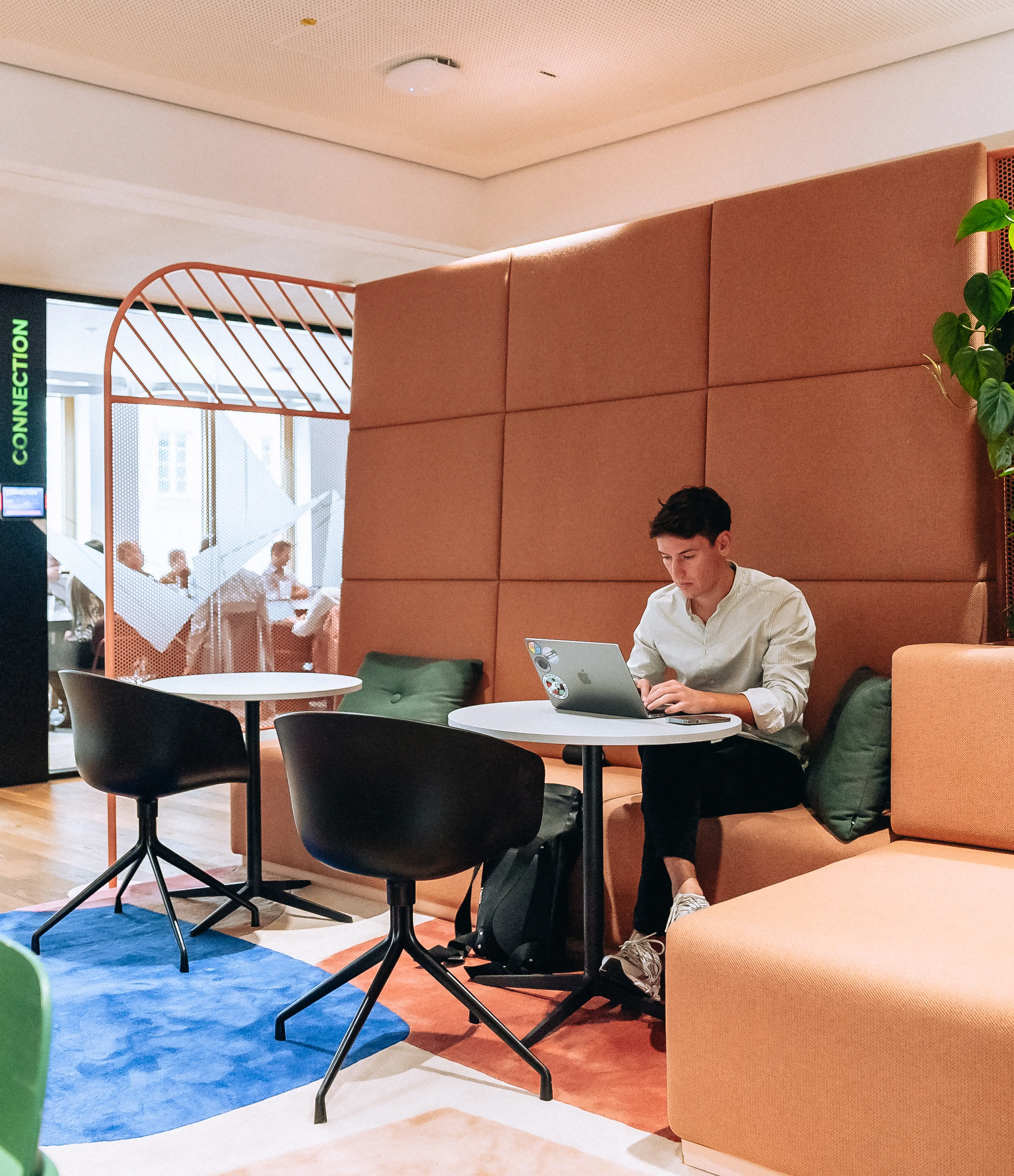 A person sits alone on an orange couch with a laptop in a brightly lit office lounge, with others visible in a nearby meeting room.