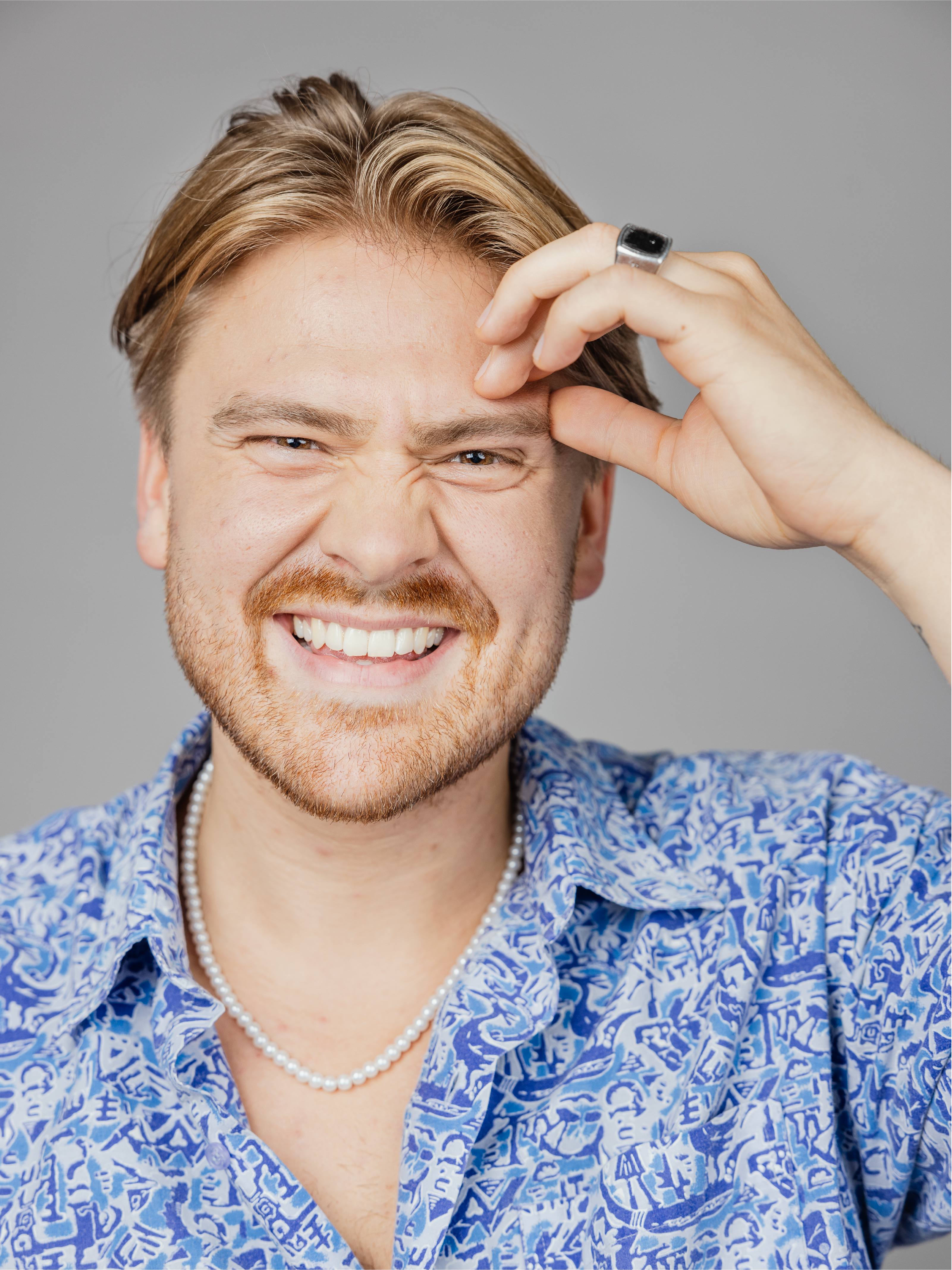 Man in blue shirt and pearl neckless, smiling and resting his hand on his forehead