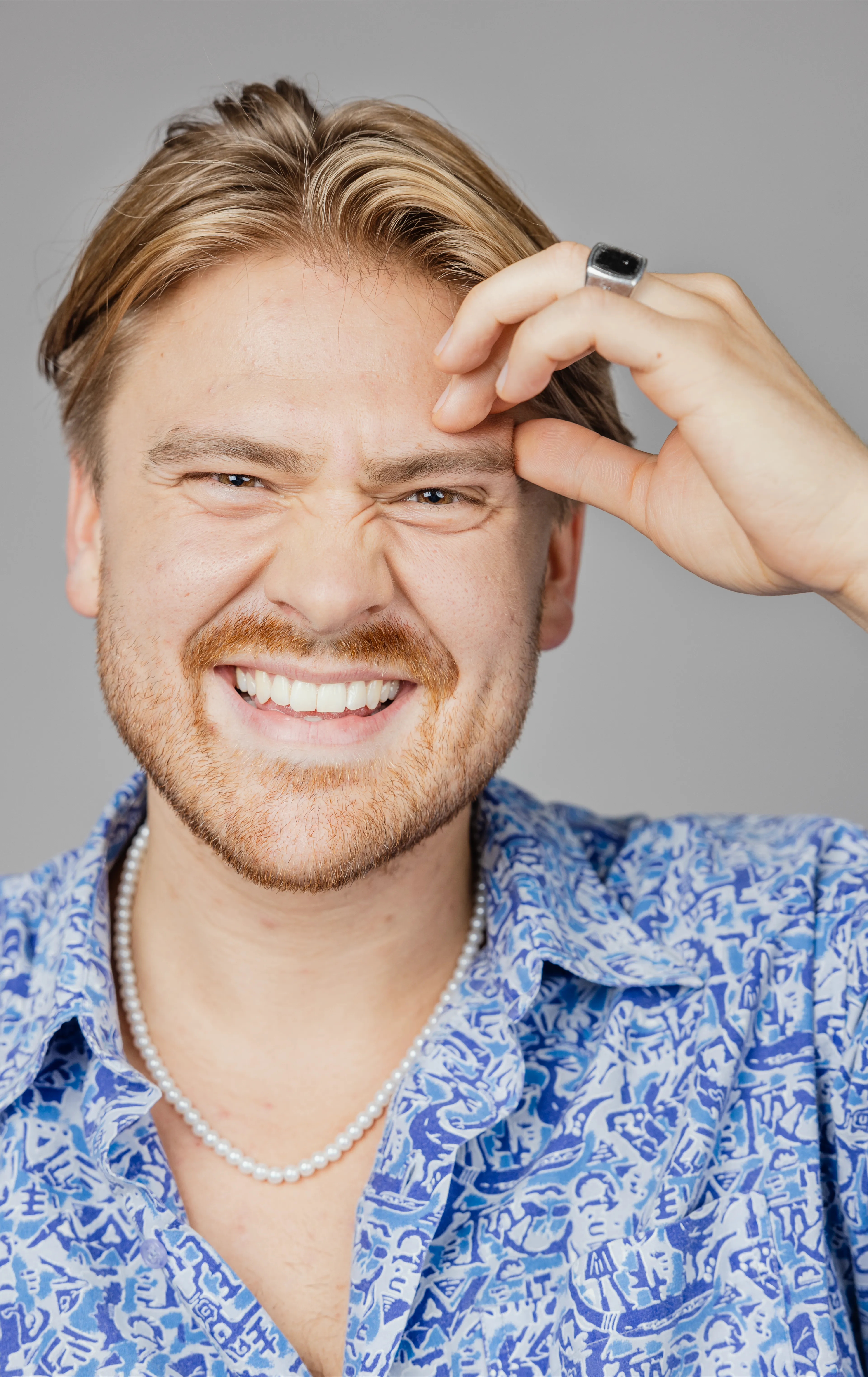 Man in blue shirt and pearl neckless, smiling and resting his hand on his forehead