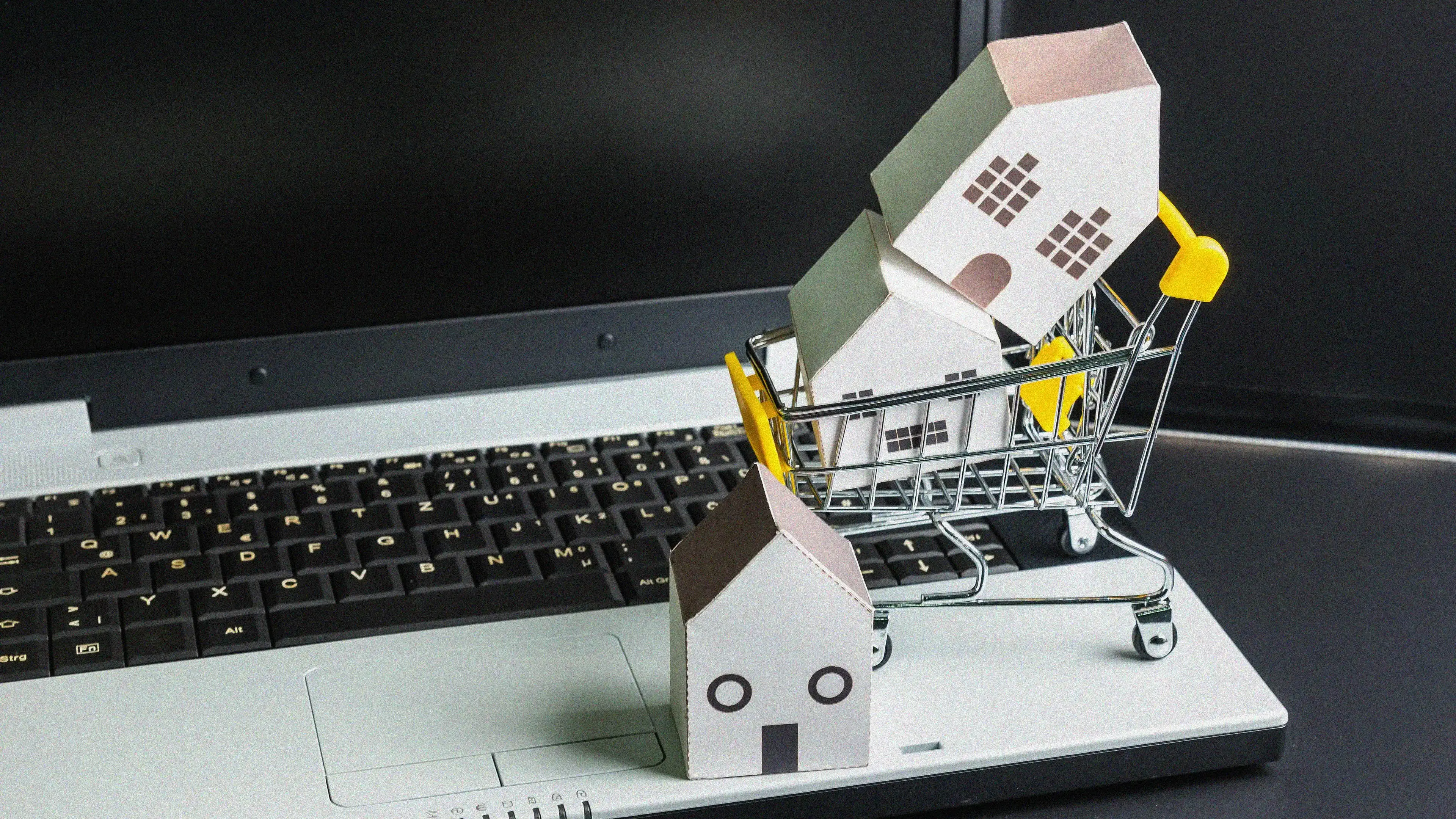 A laptop with a miniature shopping cart on top of the keyboard, containing two small cardboard house models. Another cardboard house is placed in front of the cart, also on the laptop. The image represents online real estate shopping or digital house hunting.