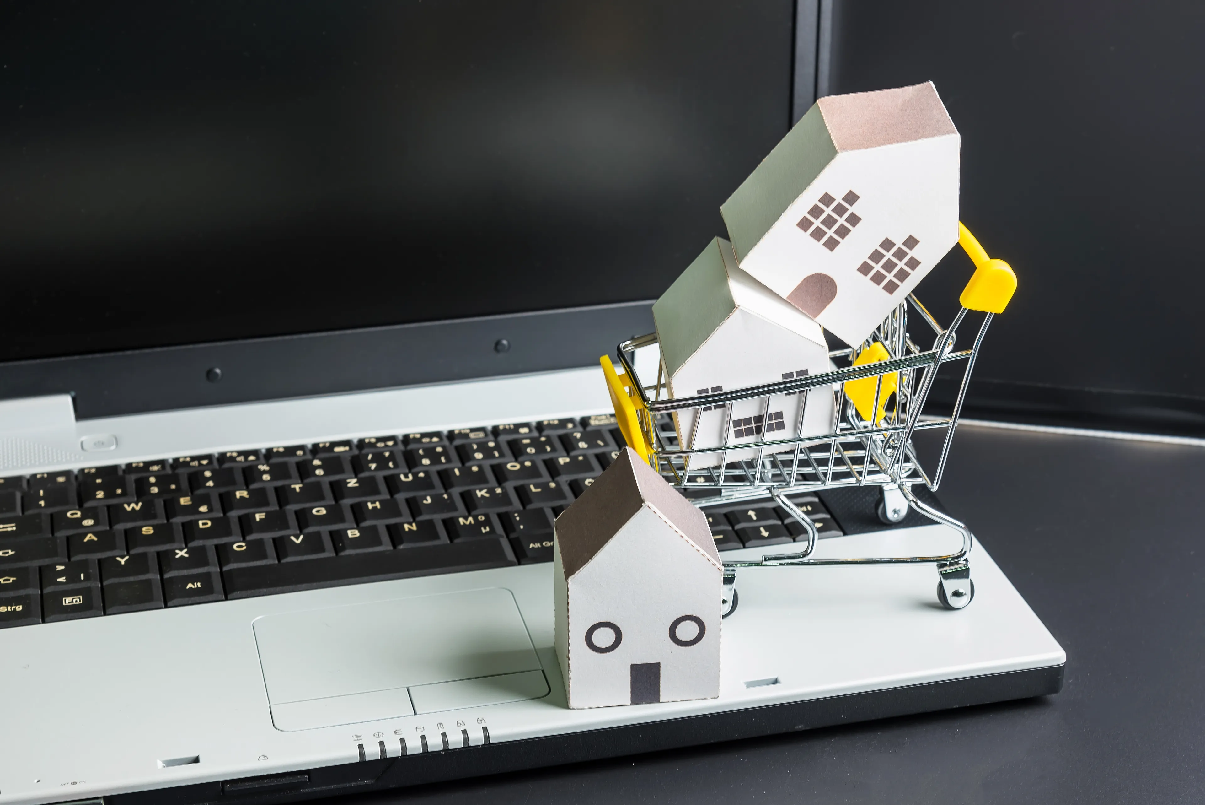 A laptop with a miniature shopping cart on top of the keyboard, containing two small cardboard house models. Another cardboard house is placed in front of the cart, also on the laptop. The image represents online real estate shopping or digital house hunting.