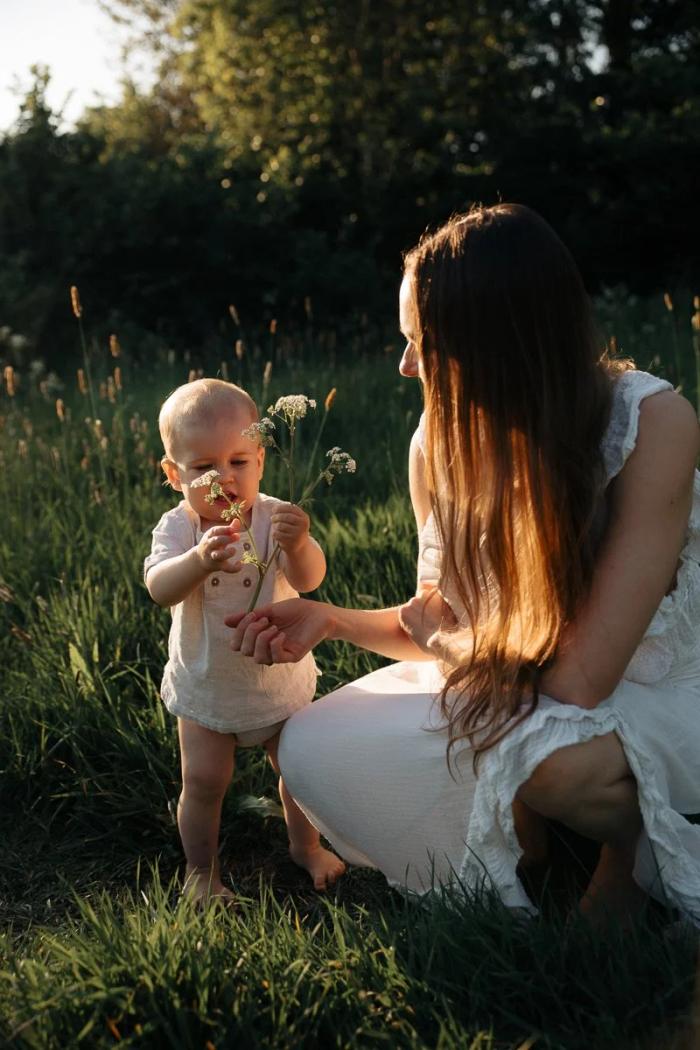 A mother crouches in long grass beside her baby, who holds a sprig of wildflowers