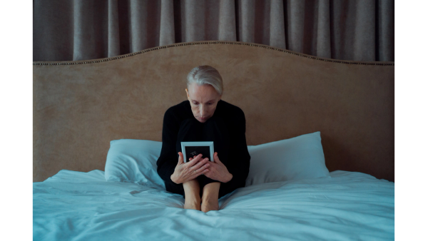 A person with grey hair sits on a bed, holding a photo frame, in a moment reflective of memory loss