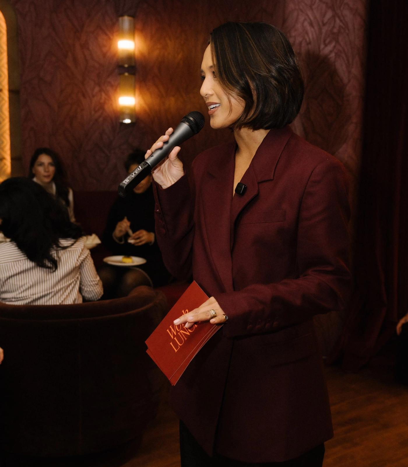 Jeanelle speaking into a microphone at a Work Lunch event, in a burgundy suit