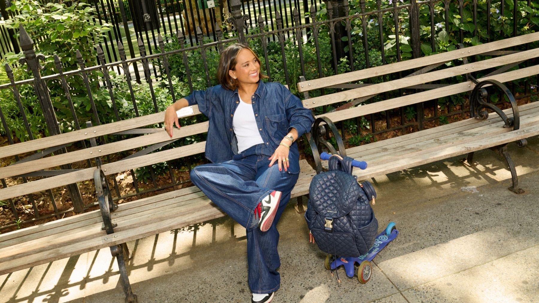 Jeanelle in denim on a park bench beside an MZ Wallace backpack and a kid’s scooter