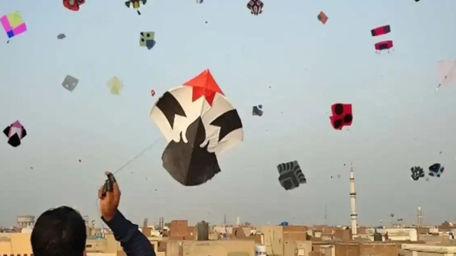 A man on a Lahore rooftop flying a black and white kite, with dozens of kites and a minaret against the sky behind him