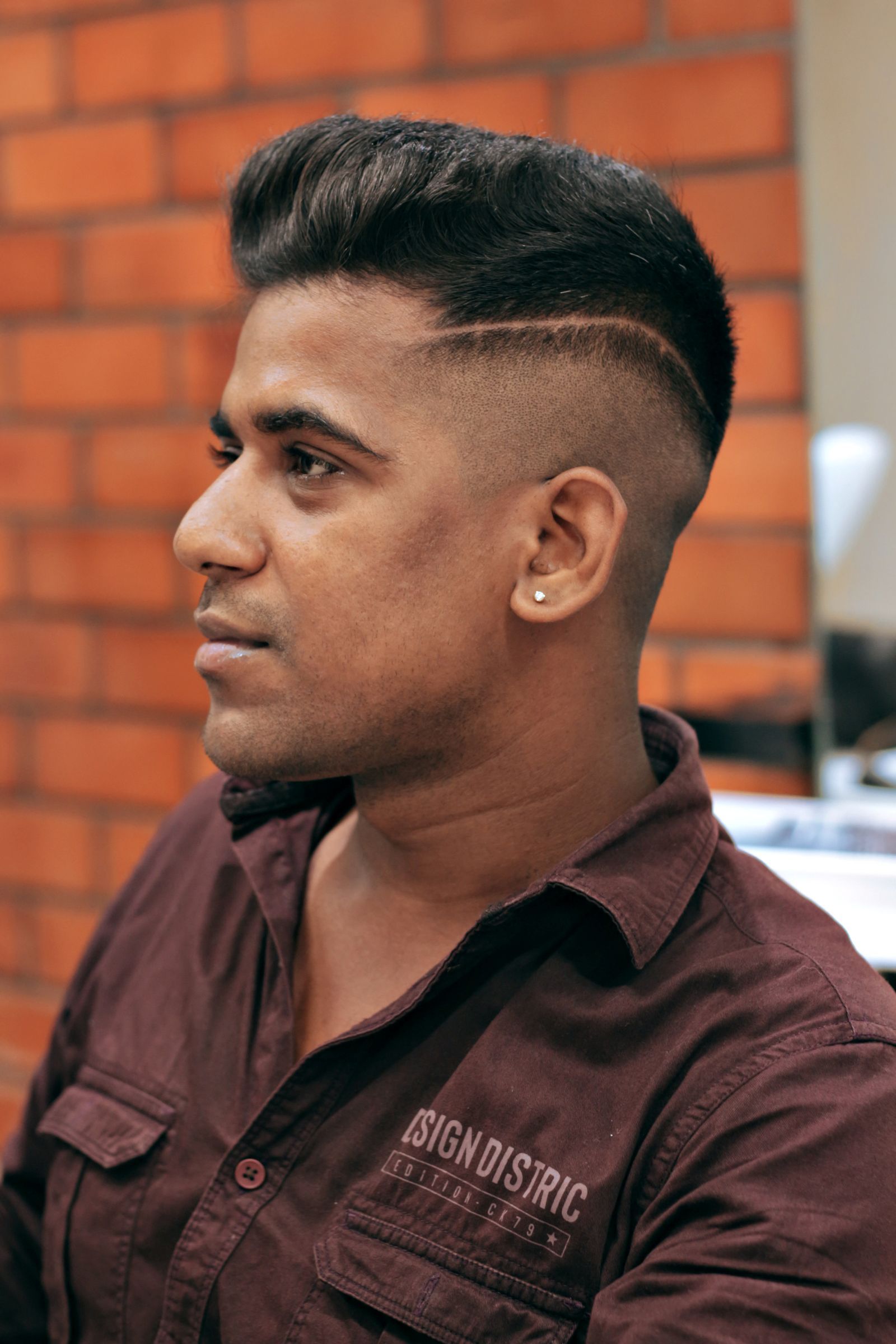 Profile of a young South Asian man with freshly styled black hair and a shaved side part, sitting in a salon against an exposed brick wall