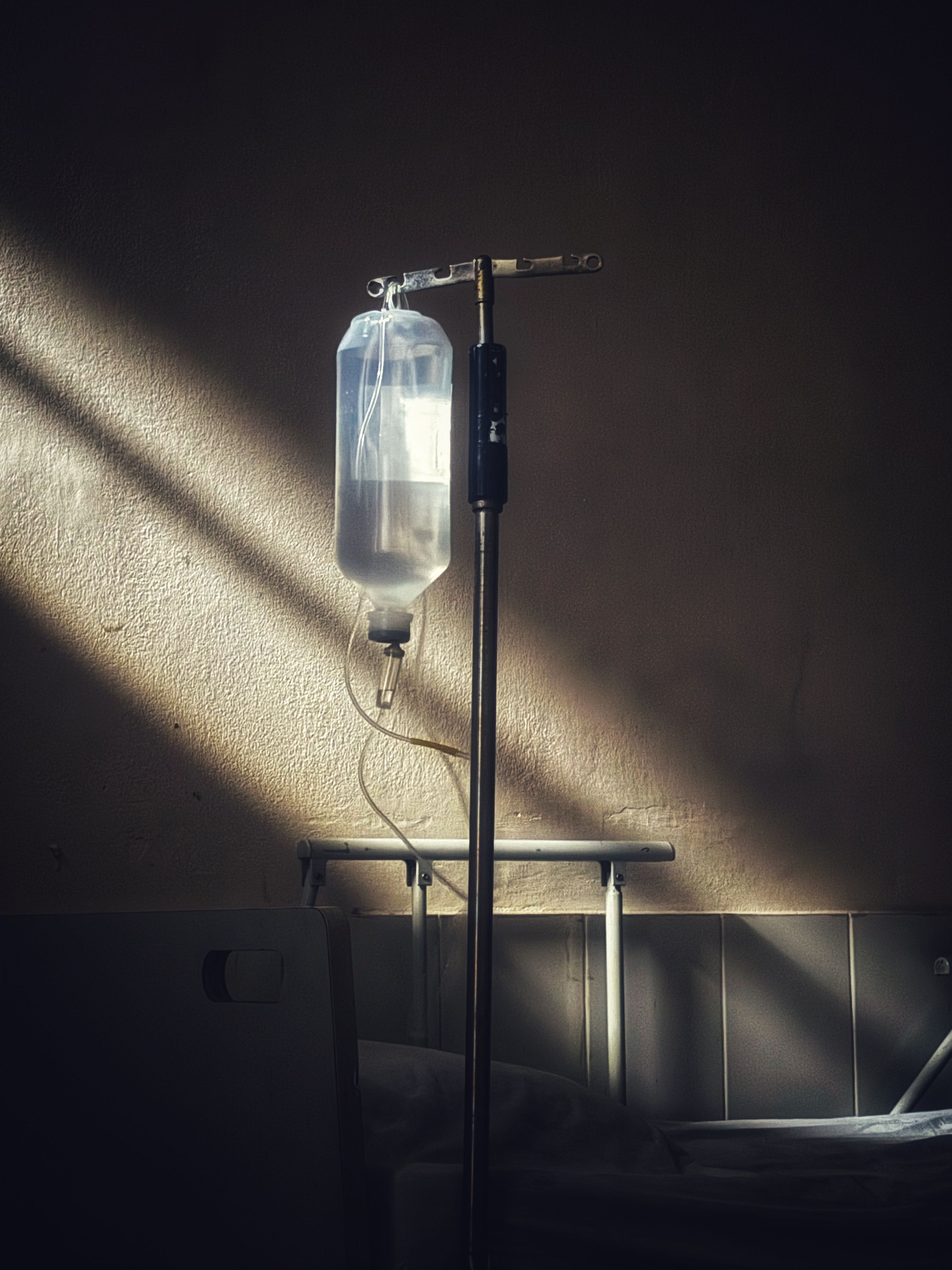Intravenous drip bottle on a stand beside a clinic bed, lit by a shaft of window light