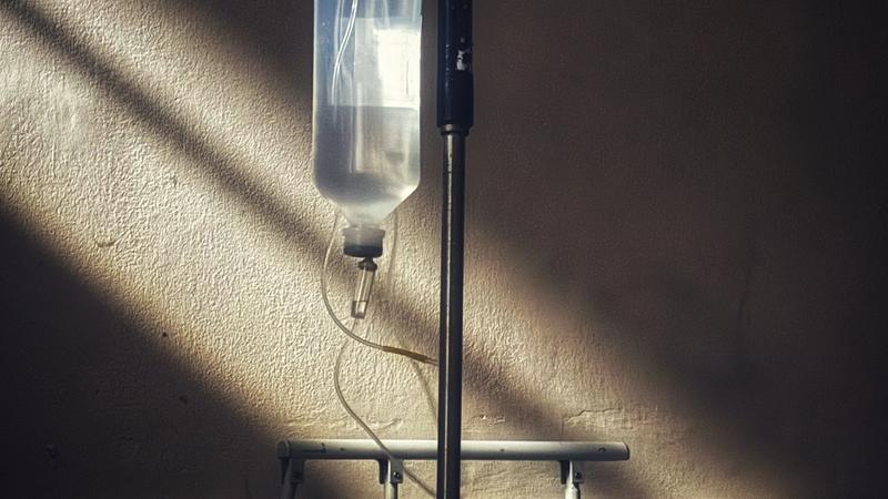 Intravenous drip bottle on a stand beside a clinic bed, lit by a shaft of window light