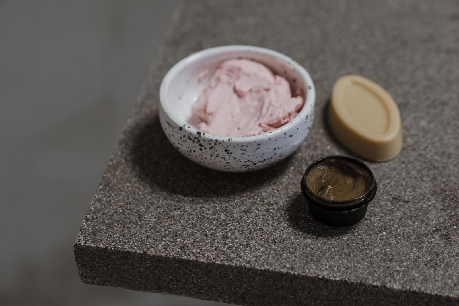 Clay face mask in a small speckled ceramic bowl beside a solid beige balm bar and a jar of amber gel on a grey stone surface