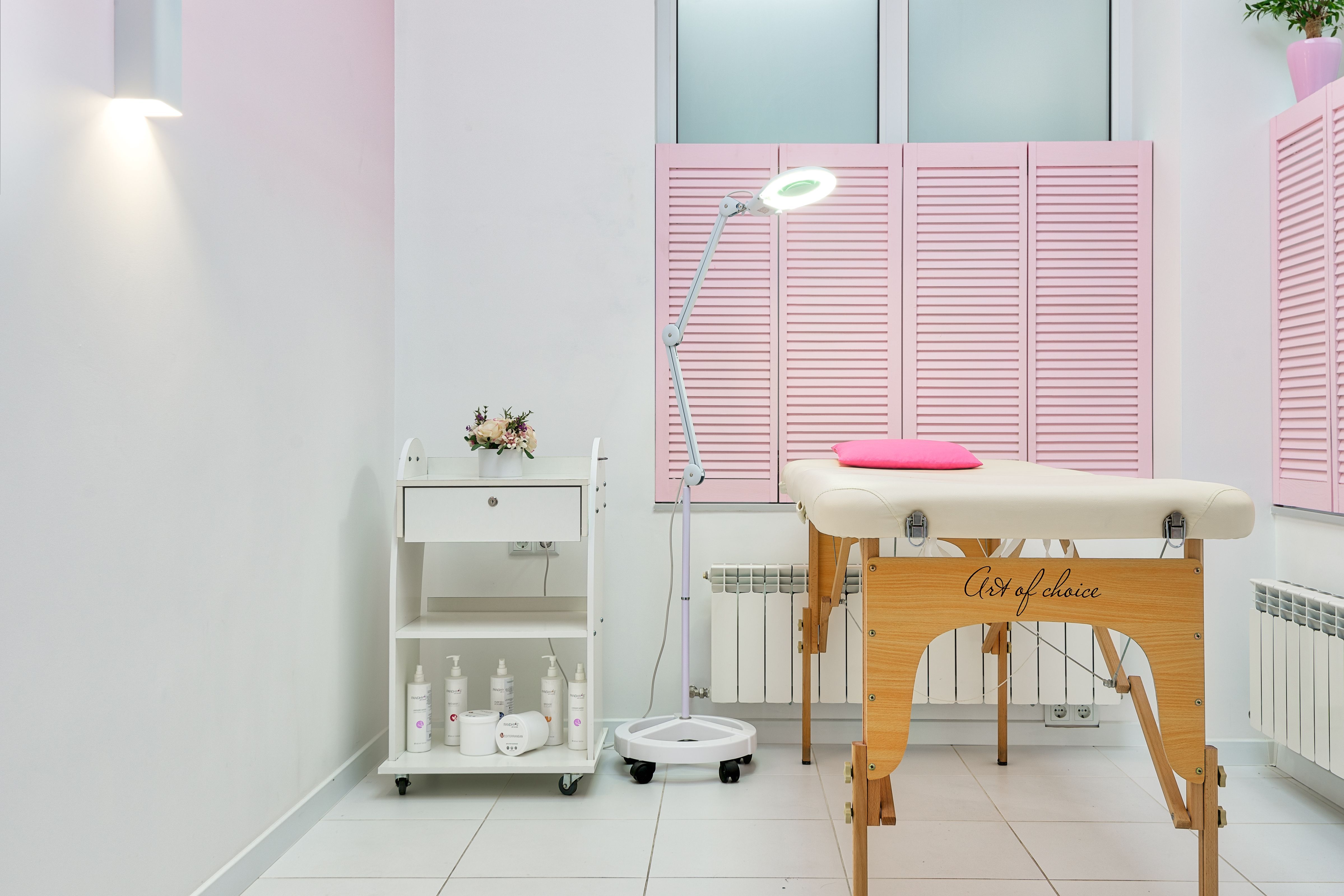 Empty salon treatment room with a cream facial bed, a magnifying ring light on a stand and a trolley of white skincare bottles