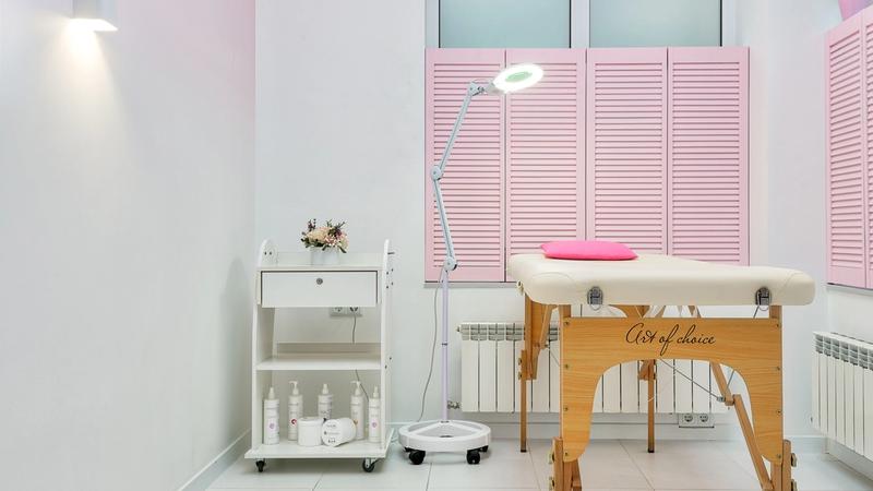 Empty salon treatment room with a cream facial bed, a magnifying ring light on a stand and a trolley of white skincare bottles