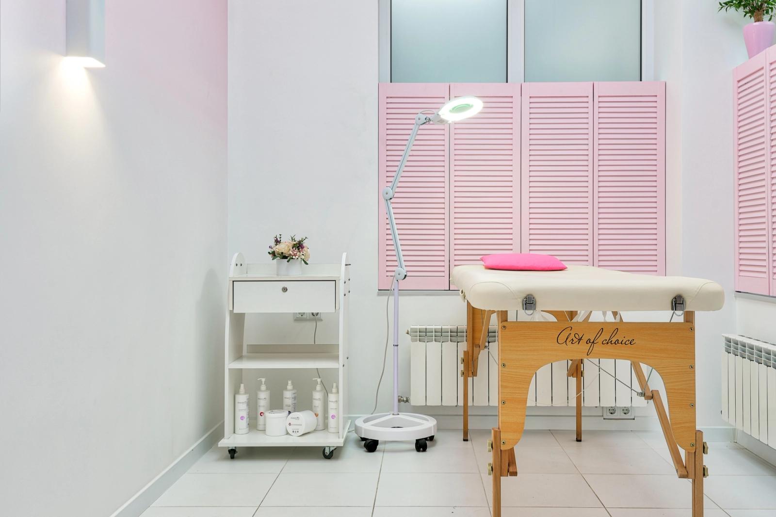 Empty salon treatment room with a cream facial bed, a magnifying ring light on a stand and a trolley of white skincare bottles