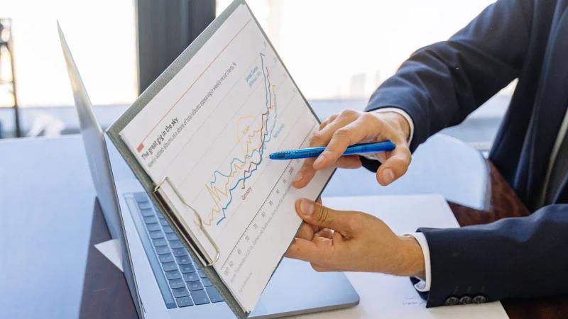 A person in a suit points with a pen at a line graph on a clipboard, with a laptop nearby.