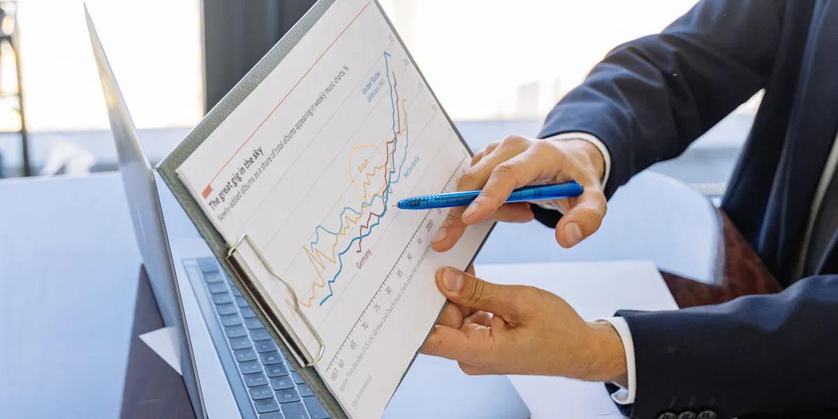 A person in a suit points with a pen at a line graph on a clipboard, with a laptop nearby.