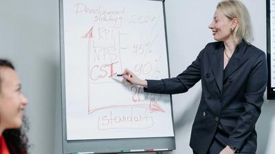 A woman in a suit smiles as she explains a development strategy written on a whiteboard to another person.
