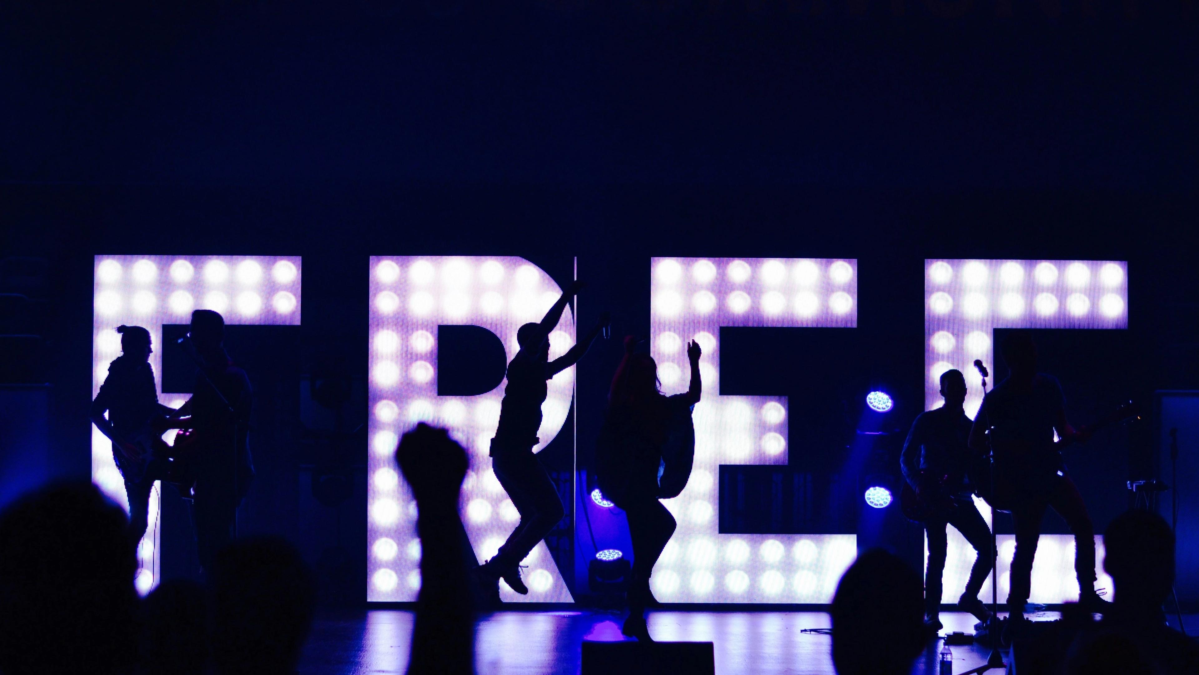 Concert with a band and dancers silhouetted against a glowing "FREE" sign.