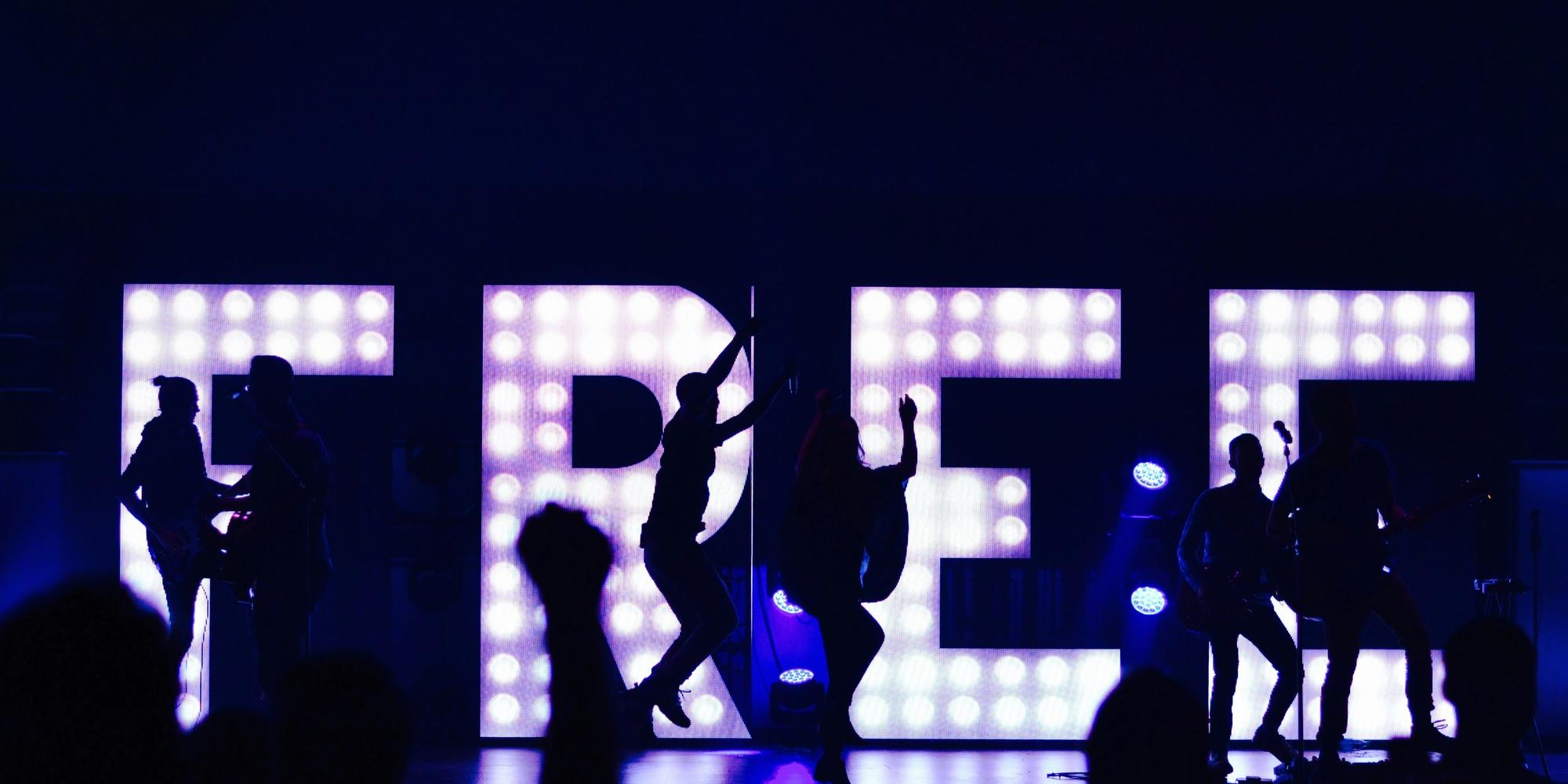 Concert with a band and dancers silhouetted against a glowing "FREE" sign.