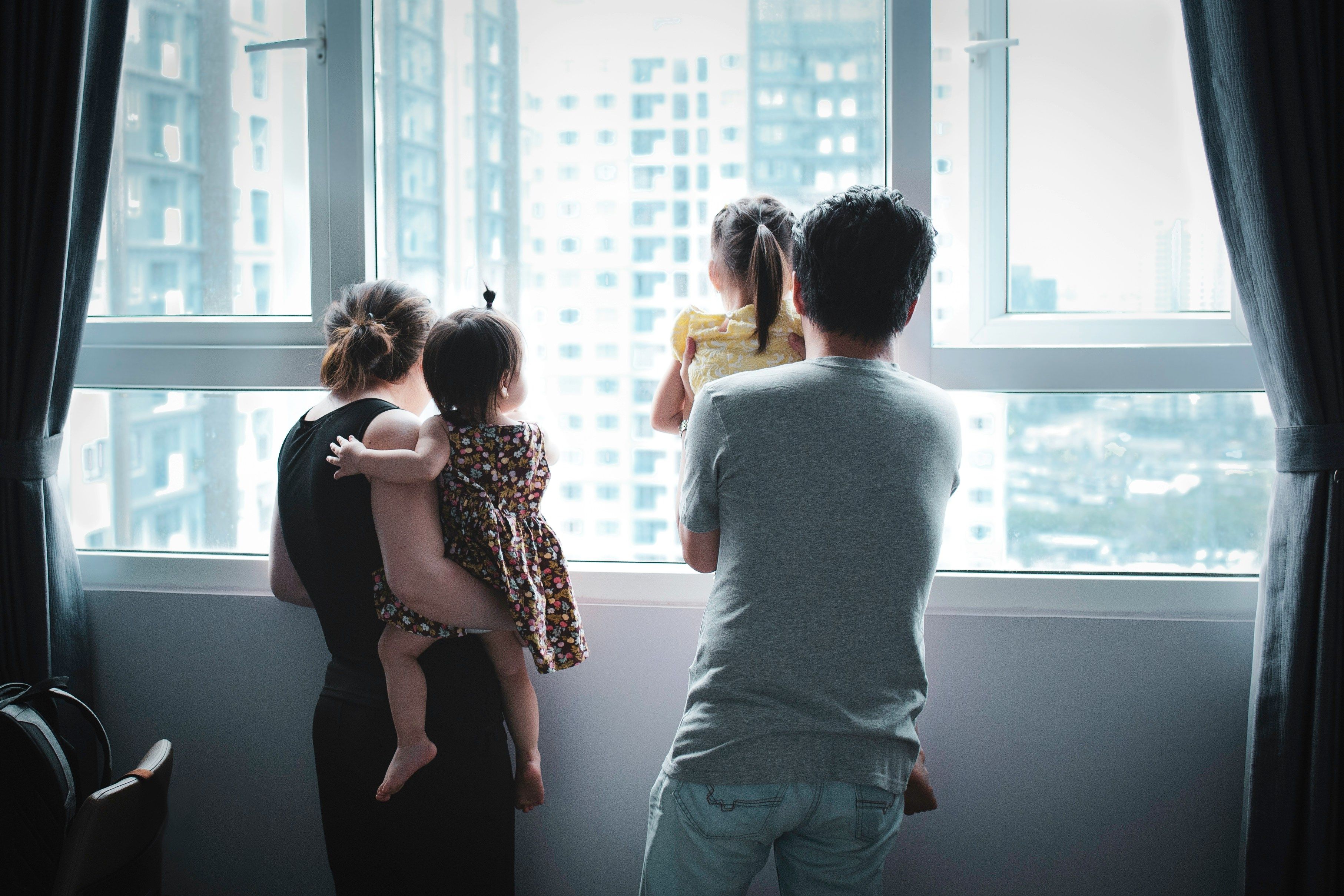 Family with two young children standing by a window and looking outside together.